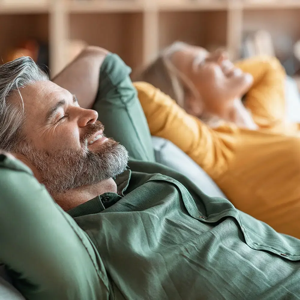 An older man with gray hair and a beard relaxing on a couch with a smile, lying back with eyes closed, in front of an older woman also lying back with eyes closed.