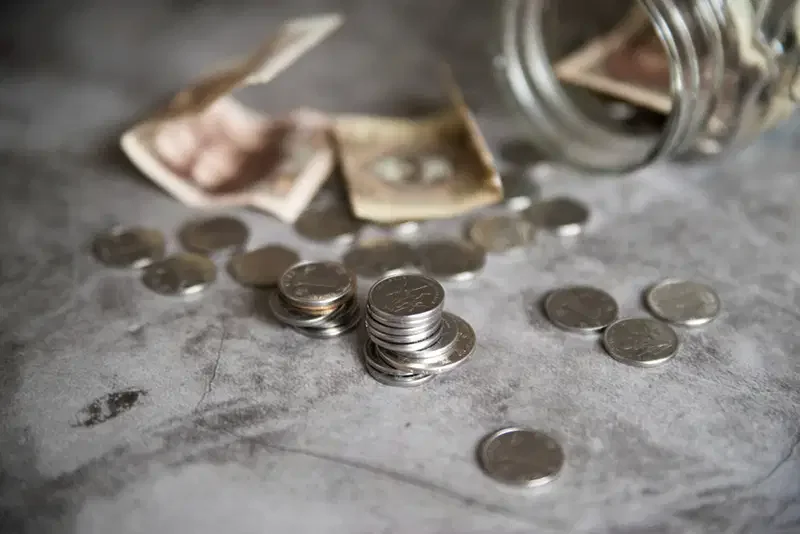 Coins and crumpled dollar bills spilled from a glass jar on a gray surface.