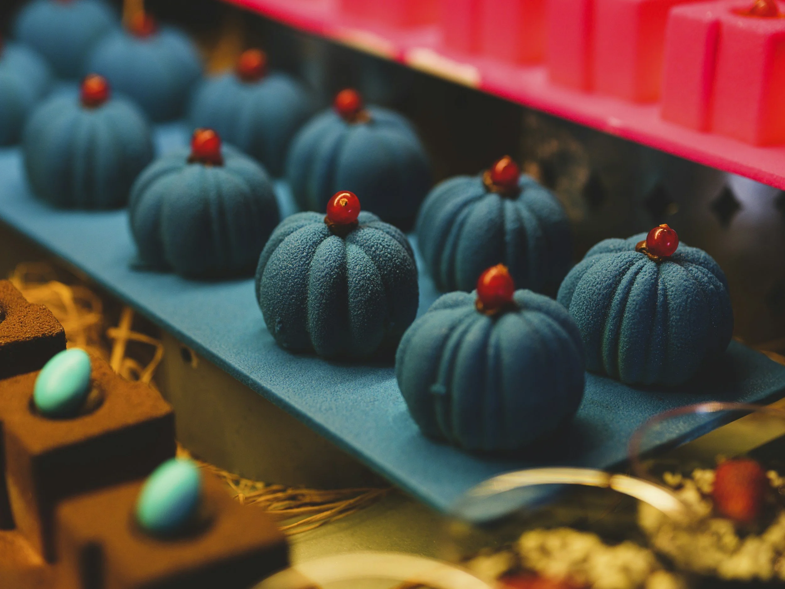 Display of blue pumpkin-shaped desserts topped with red berries, arranged on a blue tray at a bakery or dessert shop.