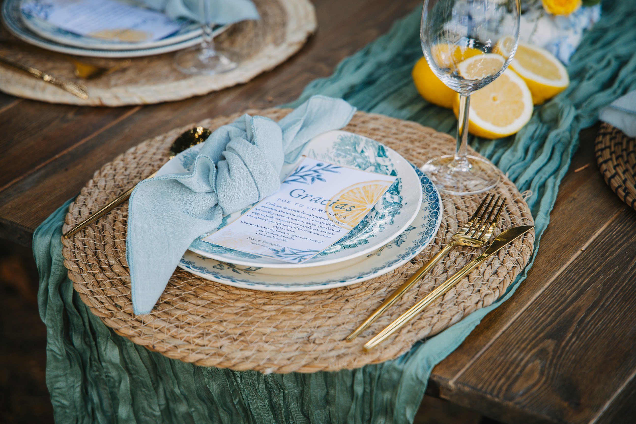 Set dining table with woven placemat, white and blue patterned plates, a light blue napkin, gold cutlery, a menu card with lemon design, a glass of water with lemon slices, and sliced lemons on a green cloth on a wooden table.