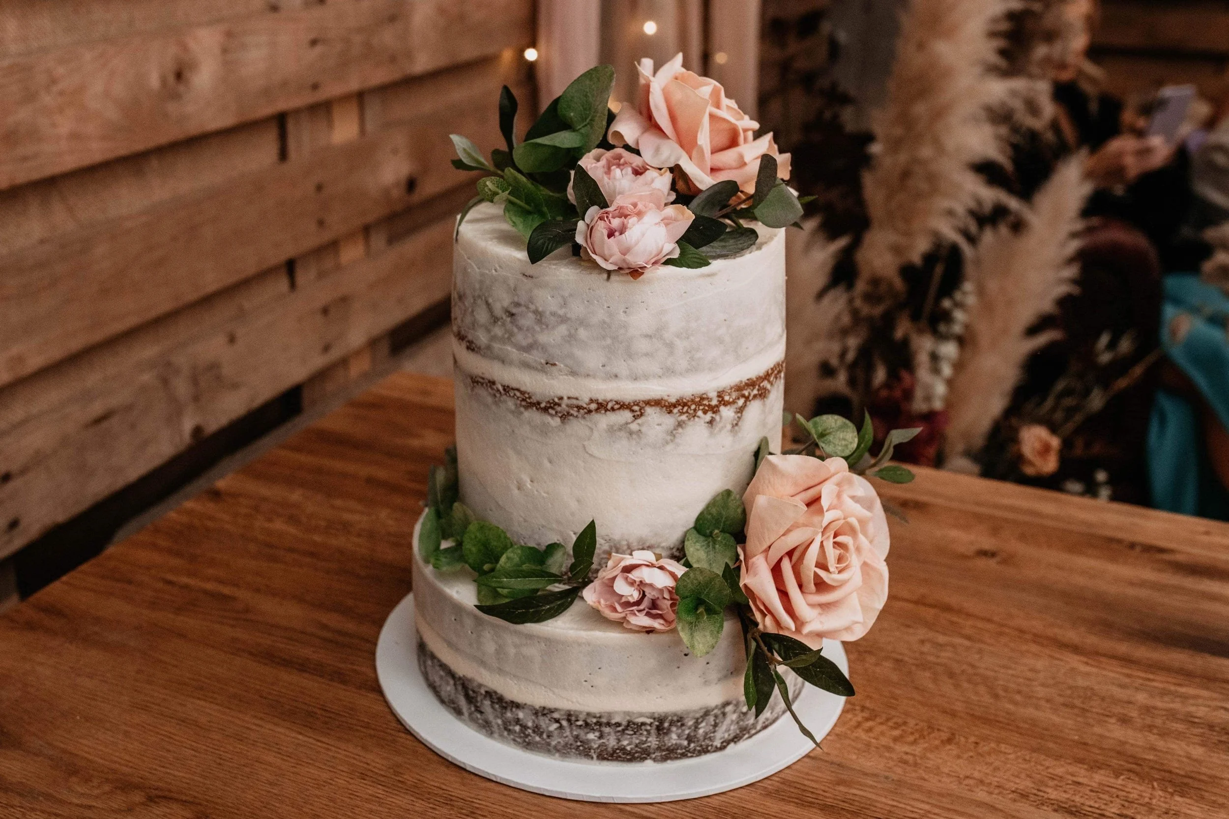 Three-tiered semi-naked wedding cake decorated with pink roses, peonies, and green leaves on a wooden table against a wood-paneled wall.