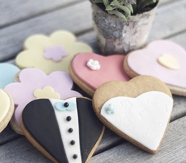 Decorated heart-shaped cookies with pastel frosting and a cloud-shaped cookie, arranged near a potted plant on a wooden surface.