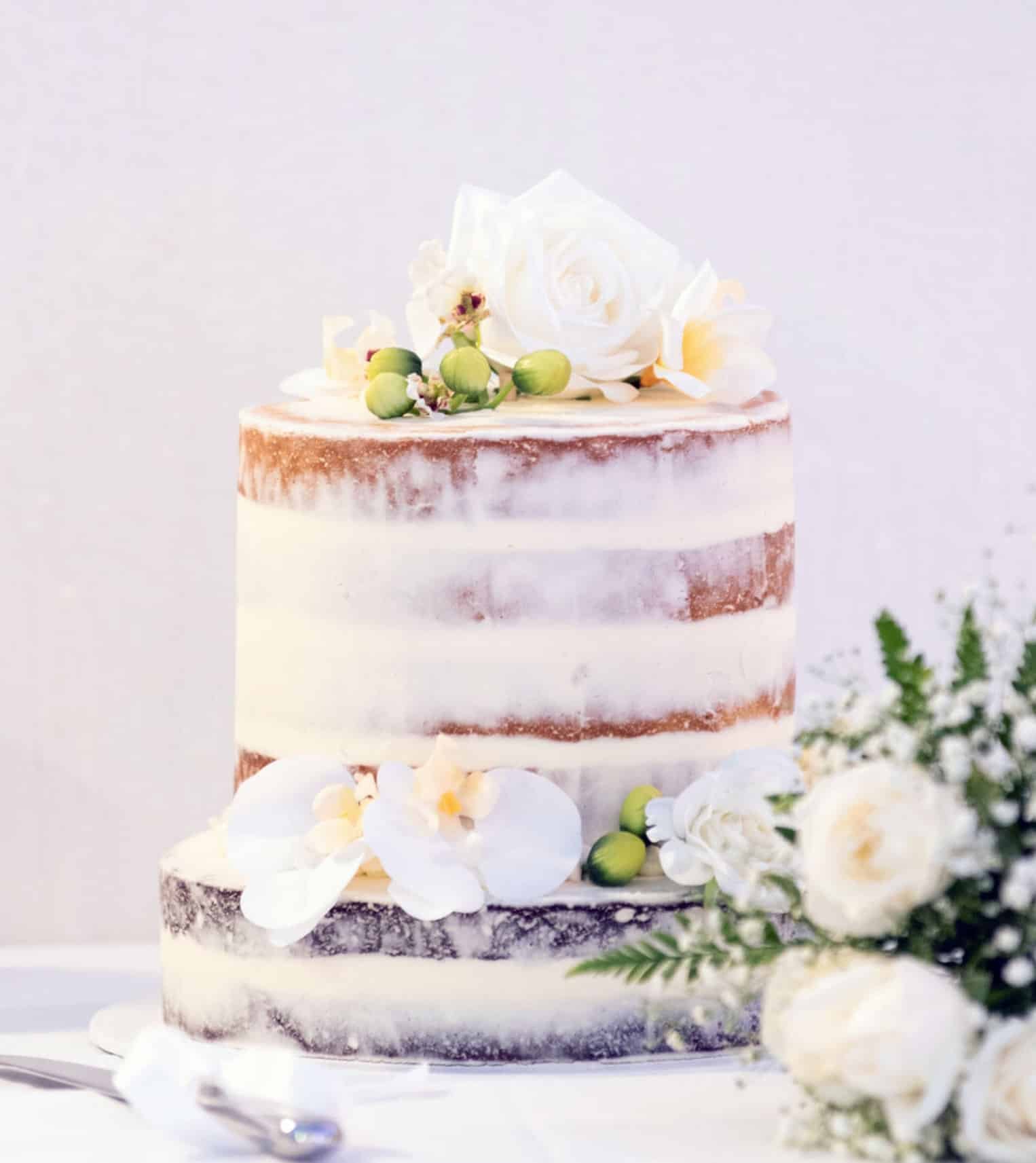 Naked wedding cake with white flowers and green berries on top, decorated with white orchids and green foliage on the side.