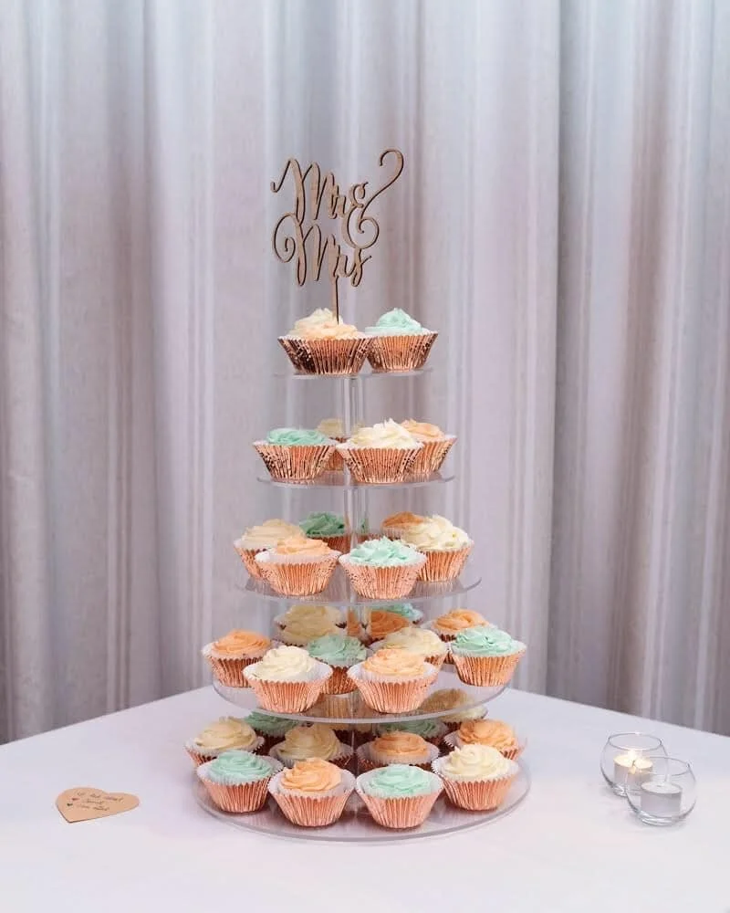 A cupcake display stand with pastel-colored cupcakes and a "Mrs & Mrs" topper, on a table with candle holders.