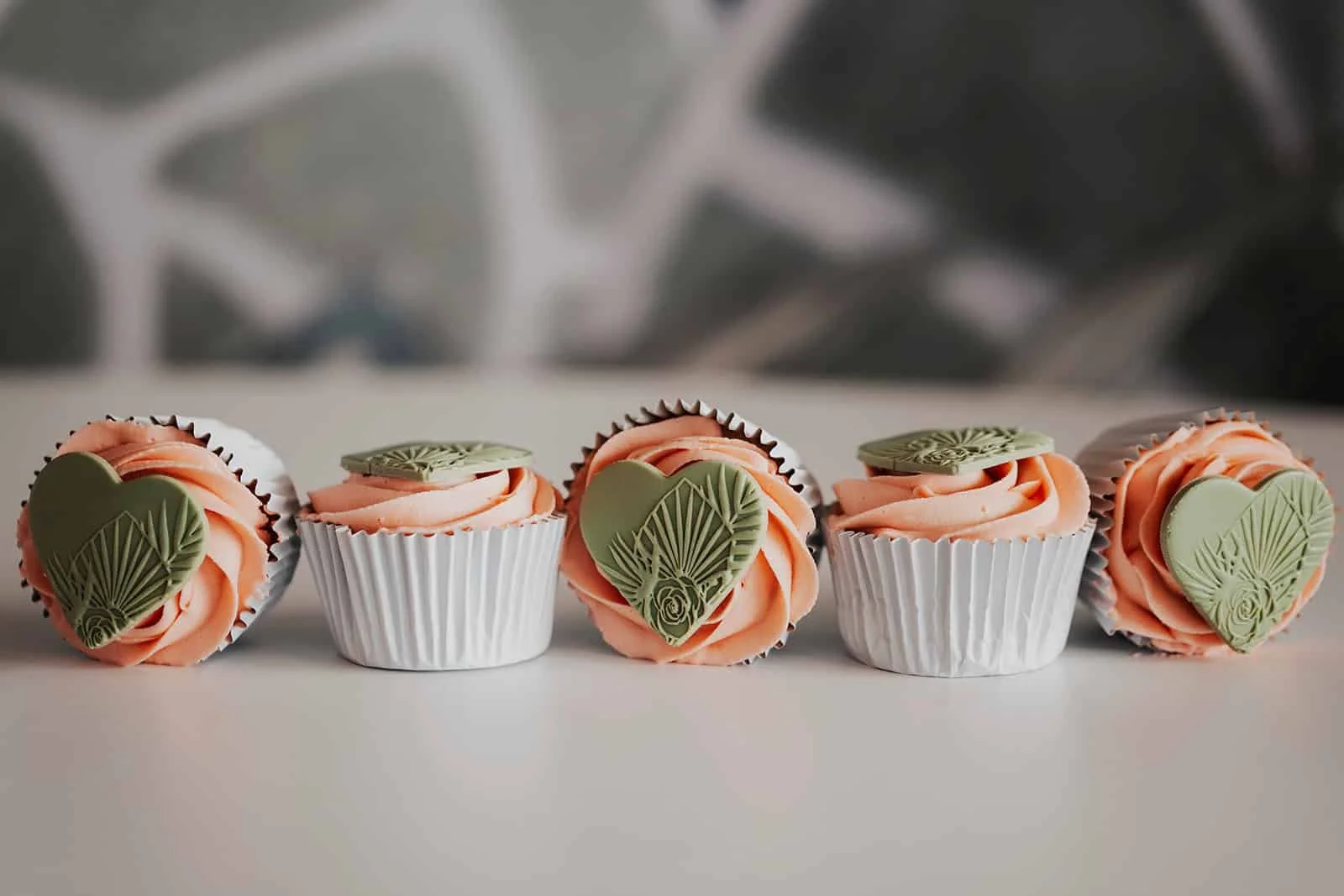 Five cupcakes with peach-colored frosting, each topped with a decorative green heart-shaped chocolate piece, arranged in a row on a white surface with a blurred background.
