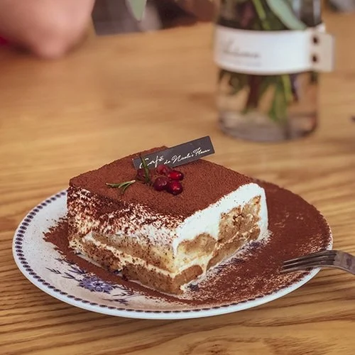 Slice of tiramisu on a decorative plate with a spoon, garnished with red berries and a sprig of green herb, dusted with cocoa powder.