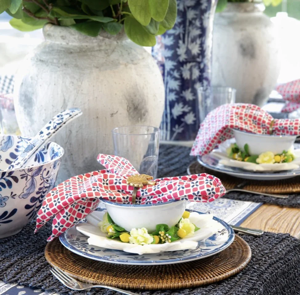 A table set with elegant blue and white dishes, floral napkins, and woven placemats, with large decorative vases in the background, suggesting a sophisticated outdoor dining setup.