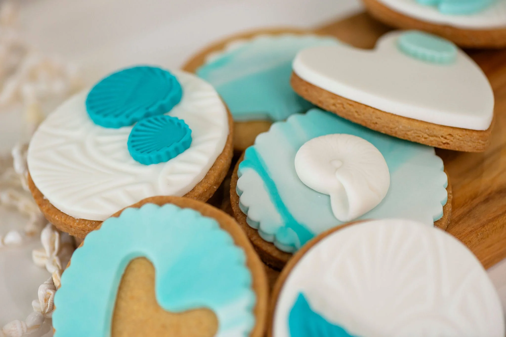 Close-up of decorated sugar cookies with ocean wave and seashell themes, featuring blue, white, and beige icing, arranged on a wooden serving board.