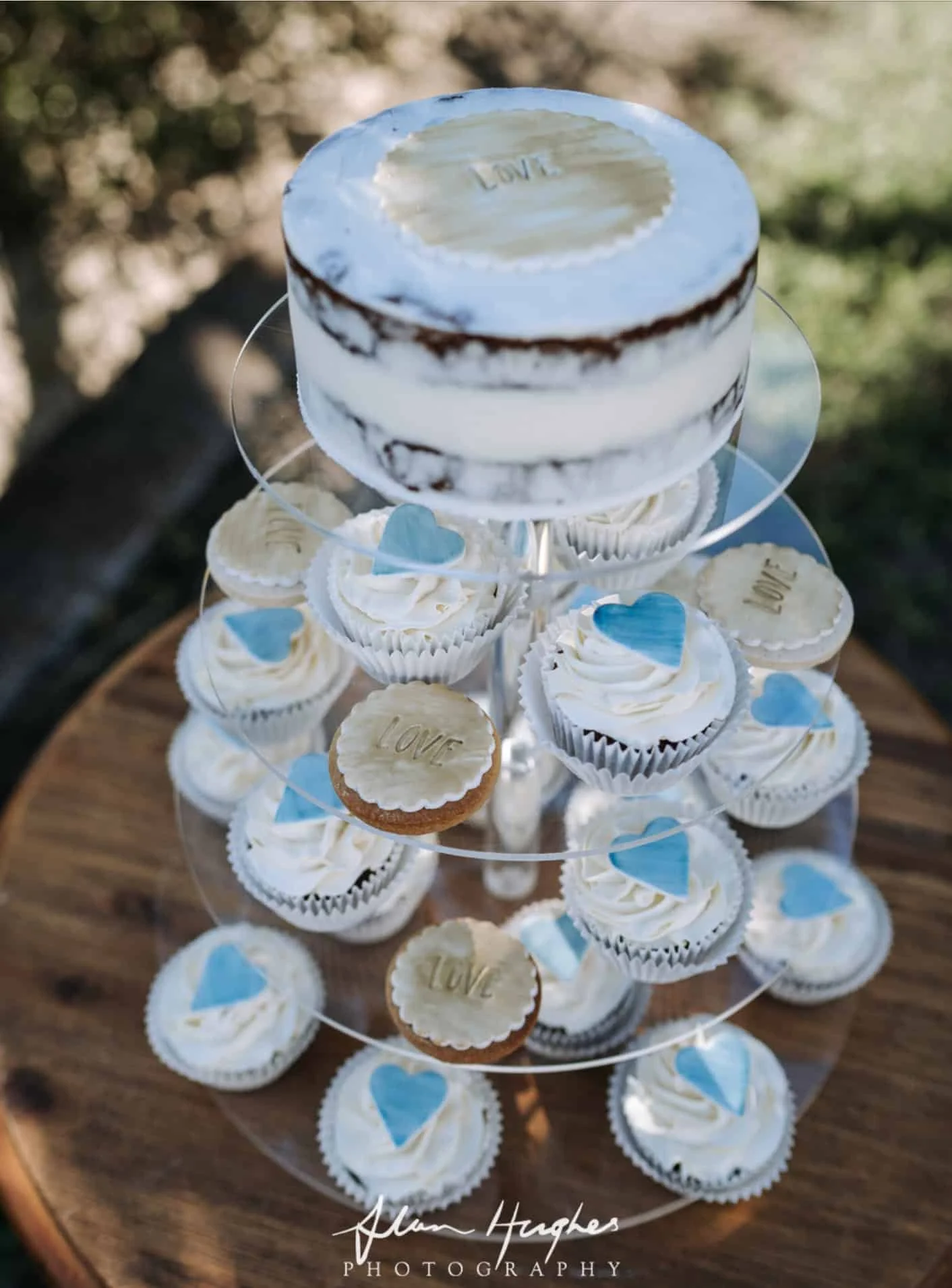 A tiered display stand with cupcakes and a small cake on top, decorated with small blue heart toppers and cookies that say 'LOVE,' set on a wooden surface outdoors.