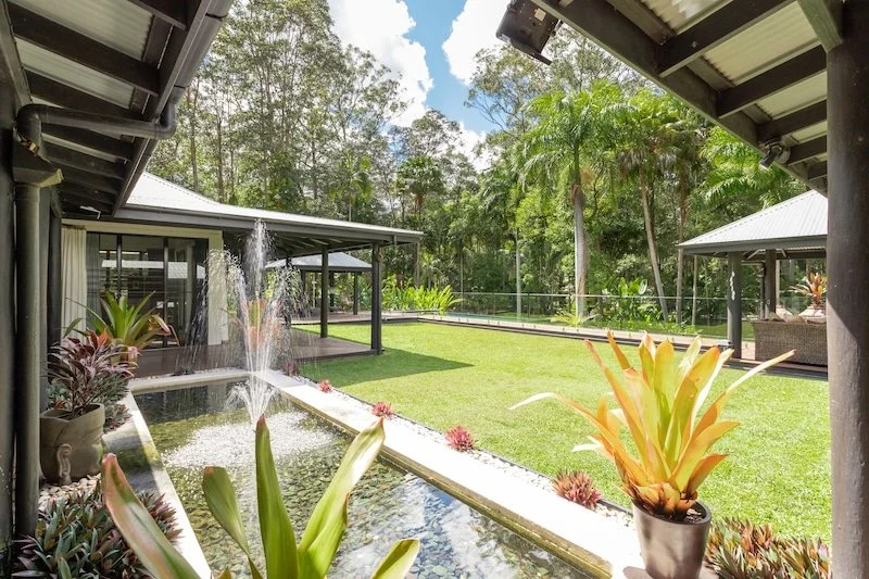 Outdoor patio with a water feature, lush green lawn, potted plants, and gazebos with seating, surrounded by trees.