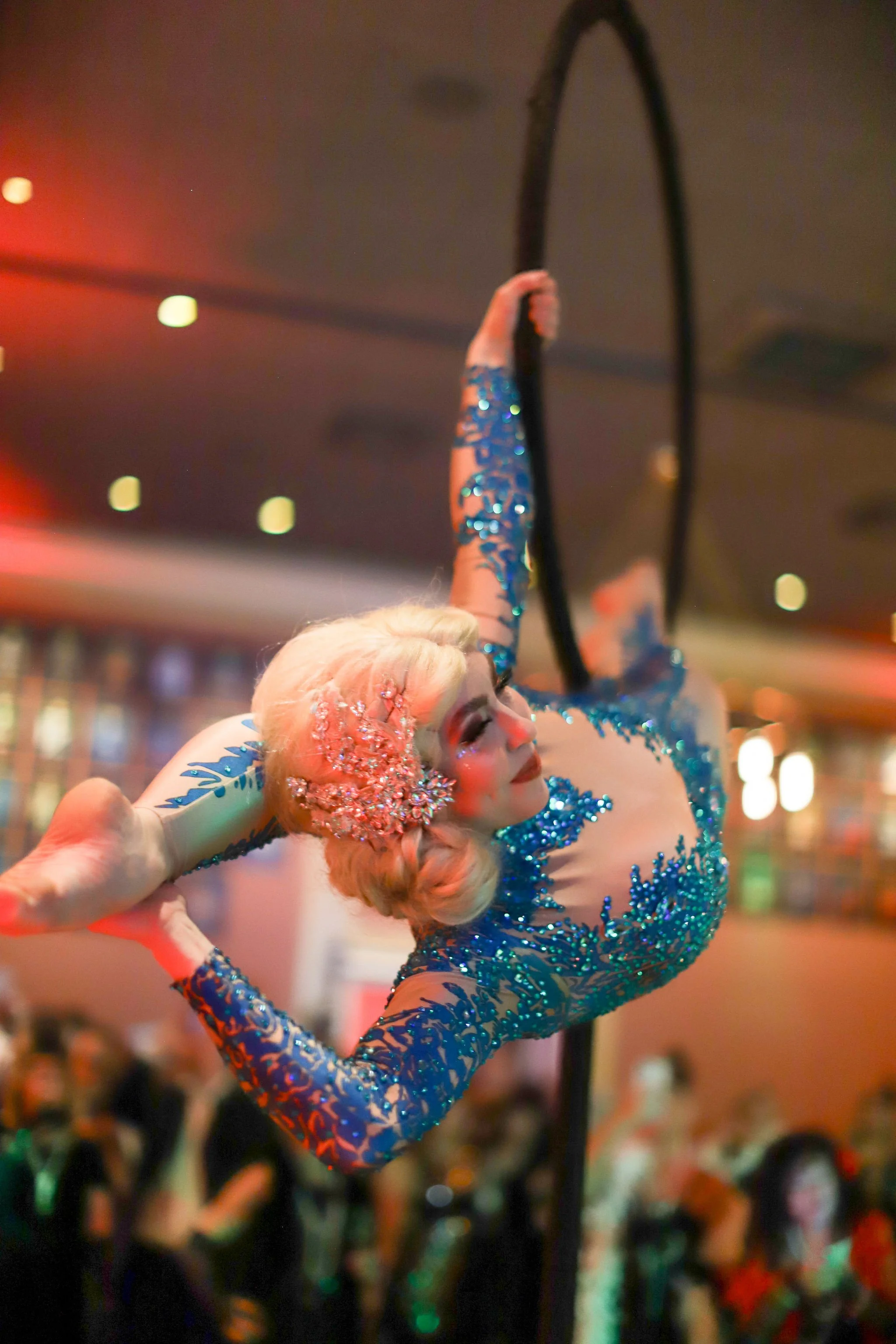 A performer in a blue, glittery costume is doing aerial hoop aerial lollipop dance at a lively event, with a crowd in the background.