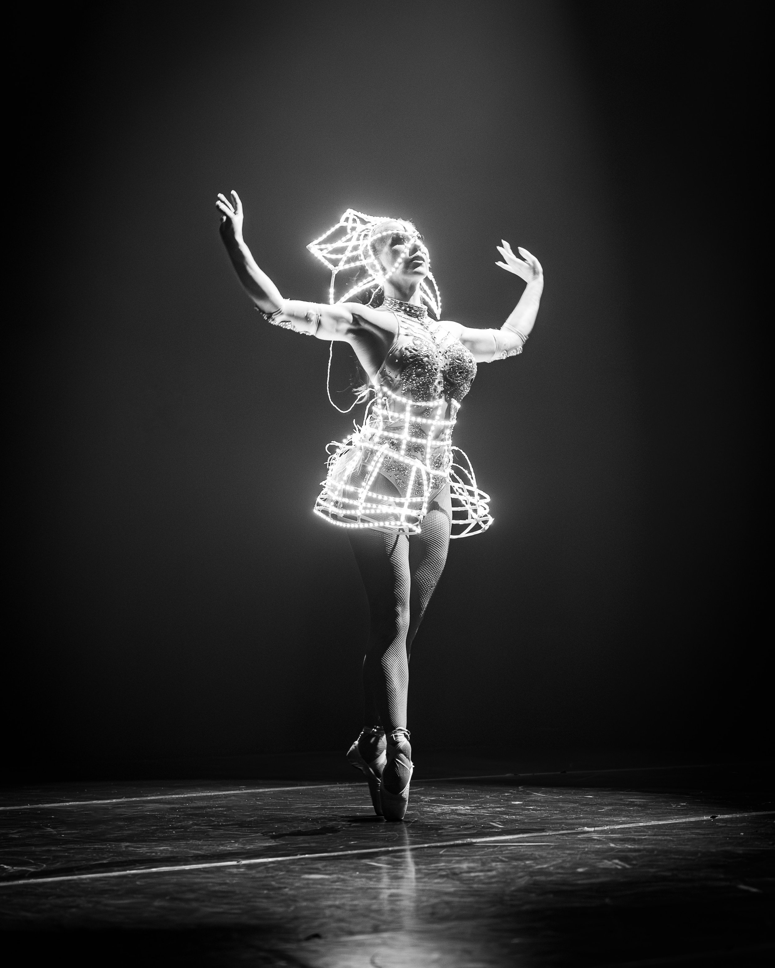 A ballet dancer performing on stage wearing a glowing LED costume, standing on toes with arms raised, in black and white.