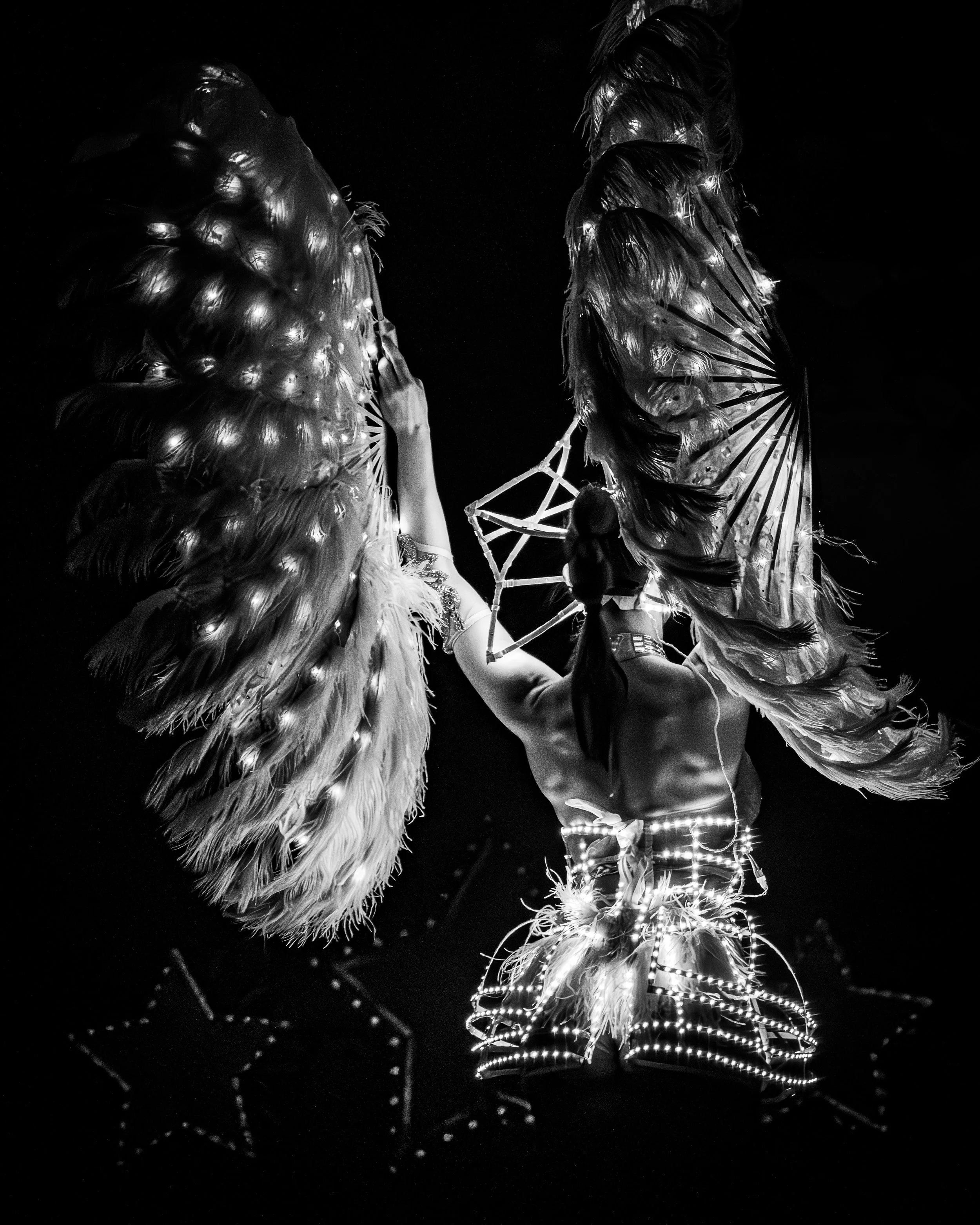 A performer from behind with large feather fans, wearing a LED costume accessories, standing against a dark background.