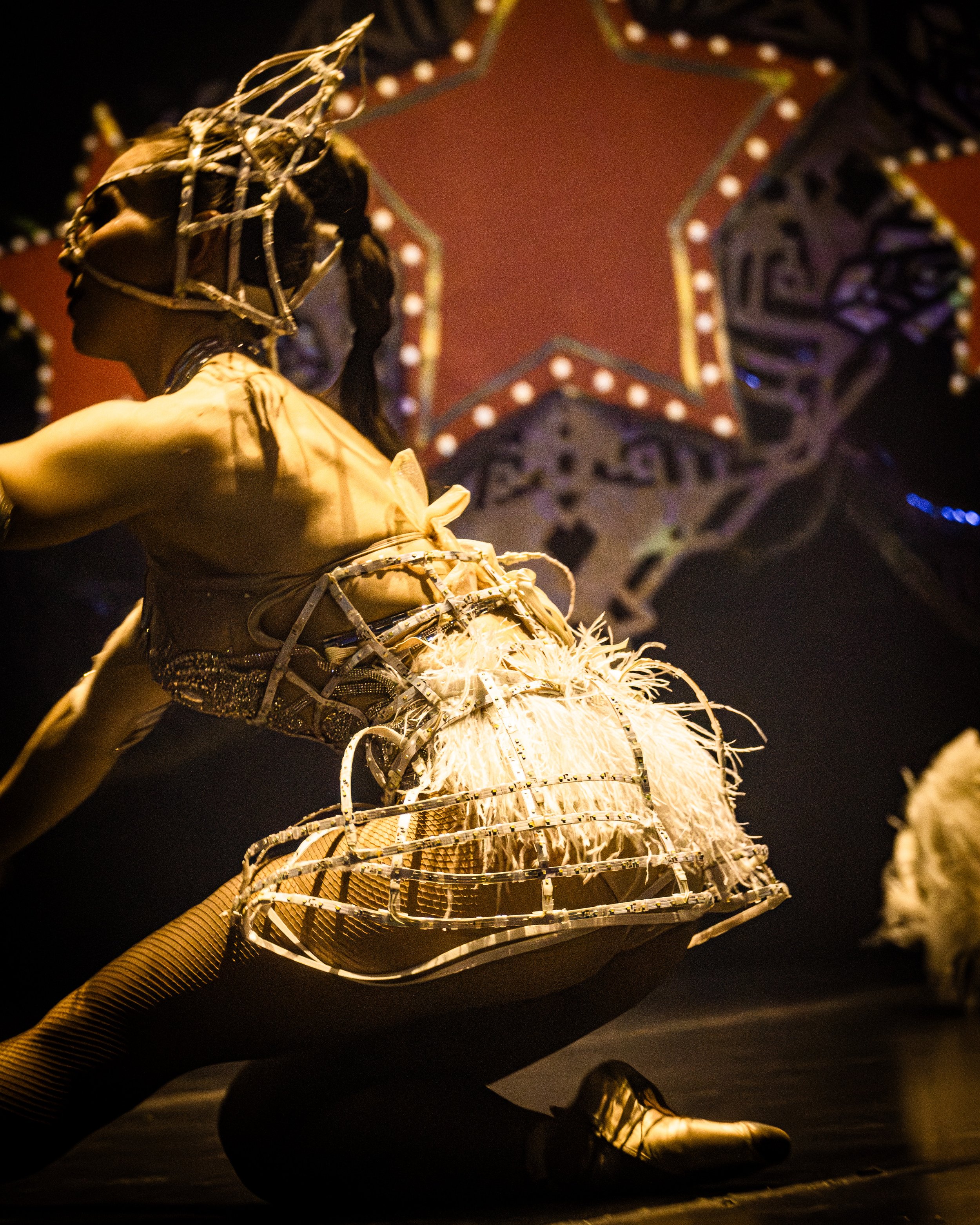A performer on stage dressed in an elaborate, metallic-looking costume with intricate details, under a large illuminated star-shaped decoration in the background.