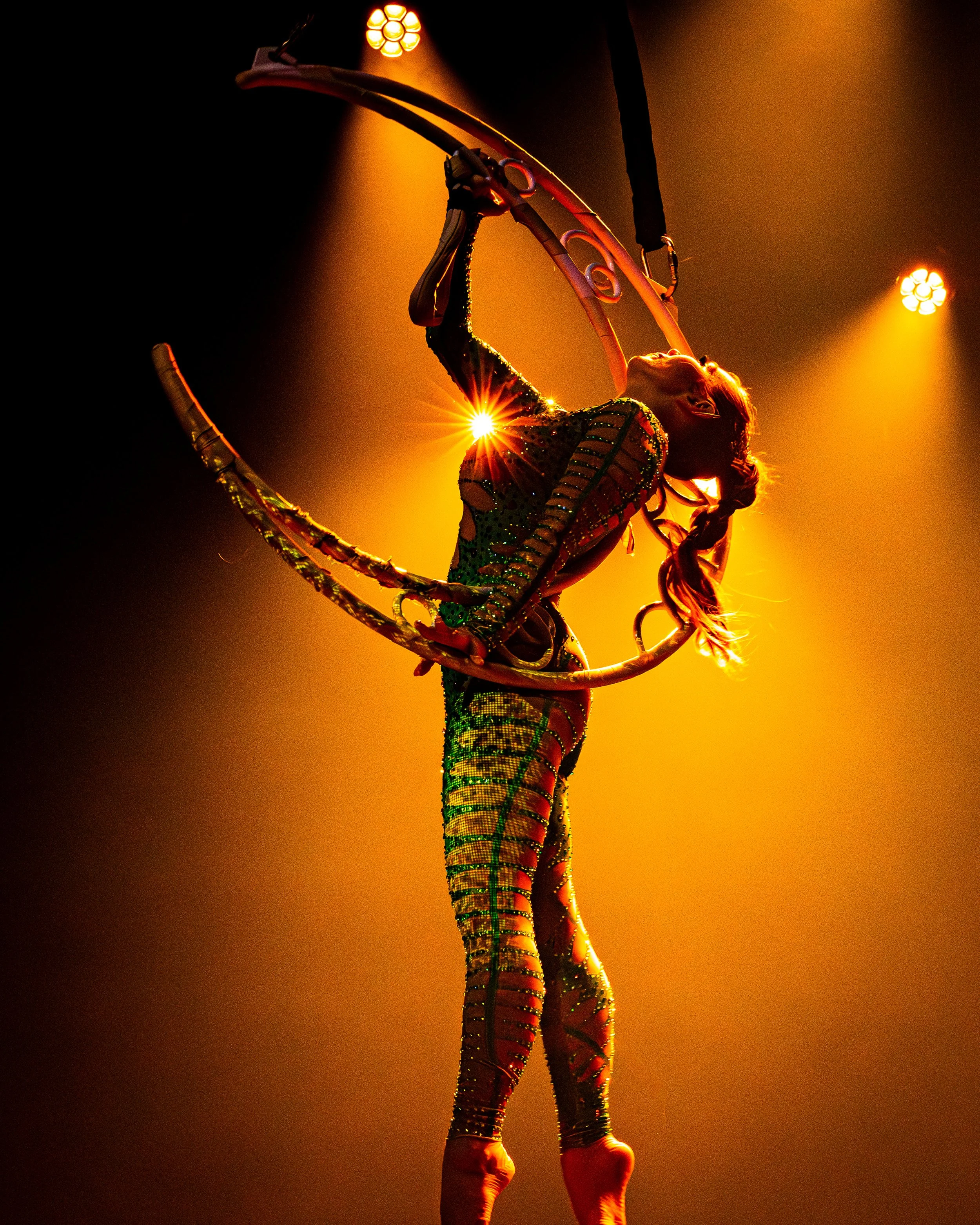 Circus performer in a glittering costume performing aerial acrobatics on a aerial moon hoop, illuminated by stage lights with a dramatic casting of shadows and bright background.