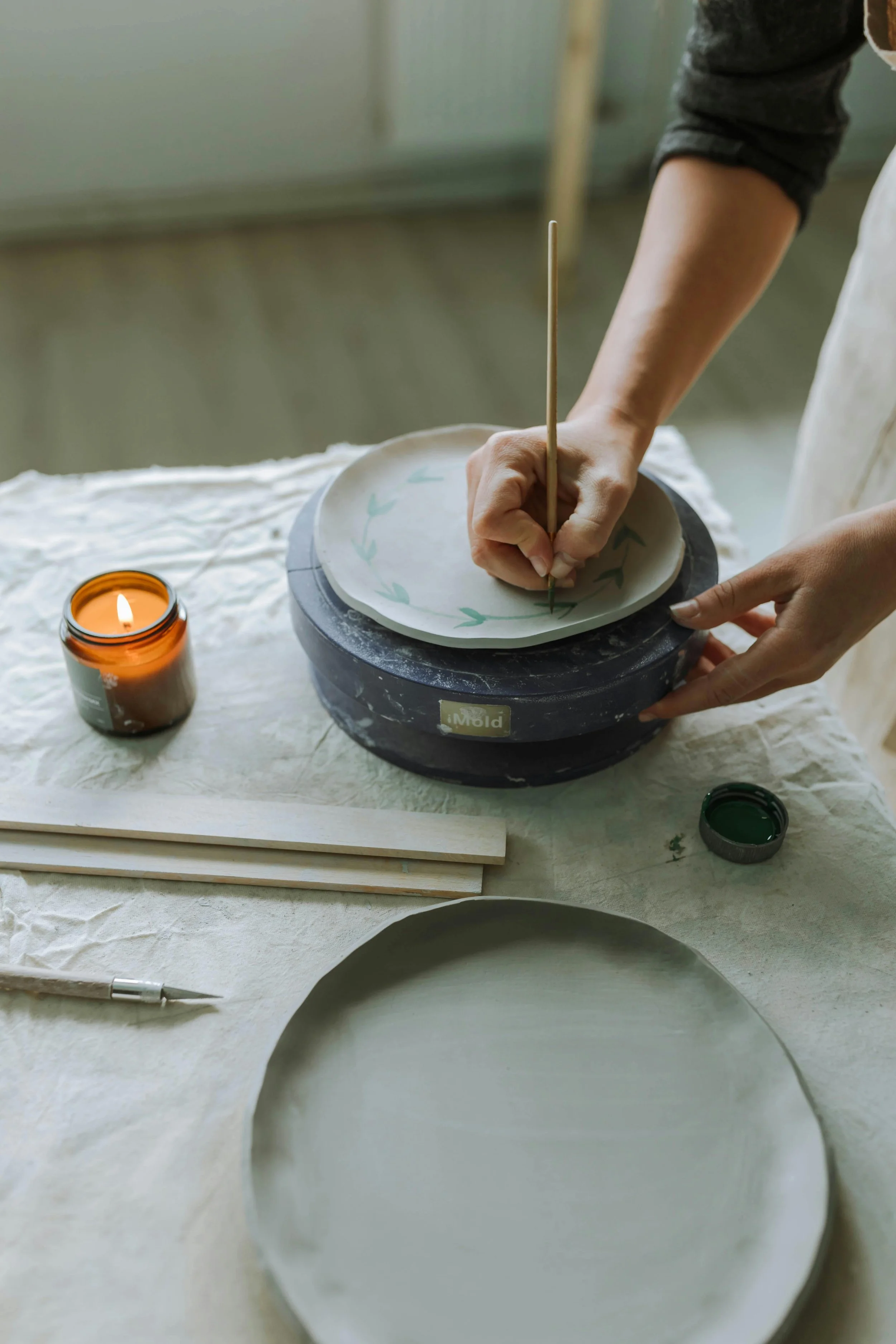 Neutral artisan workspace with a person hand-painting a clay plate on a pottery wheel, surrounded by wooden tools, a craft knife, green paint, and an amber candle. Minimal, organic, handmade aesthetic.