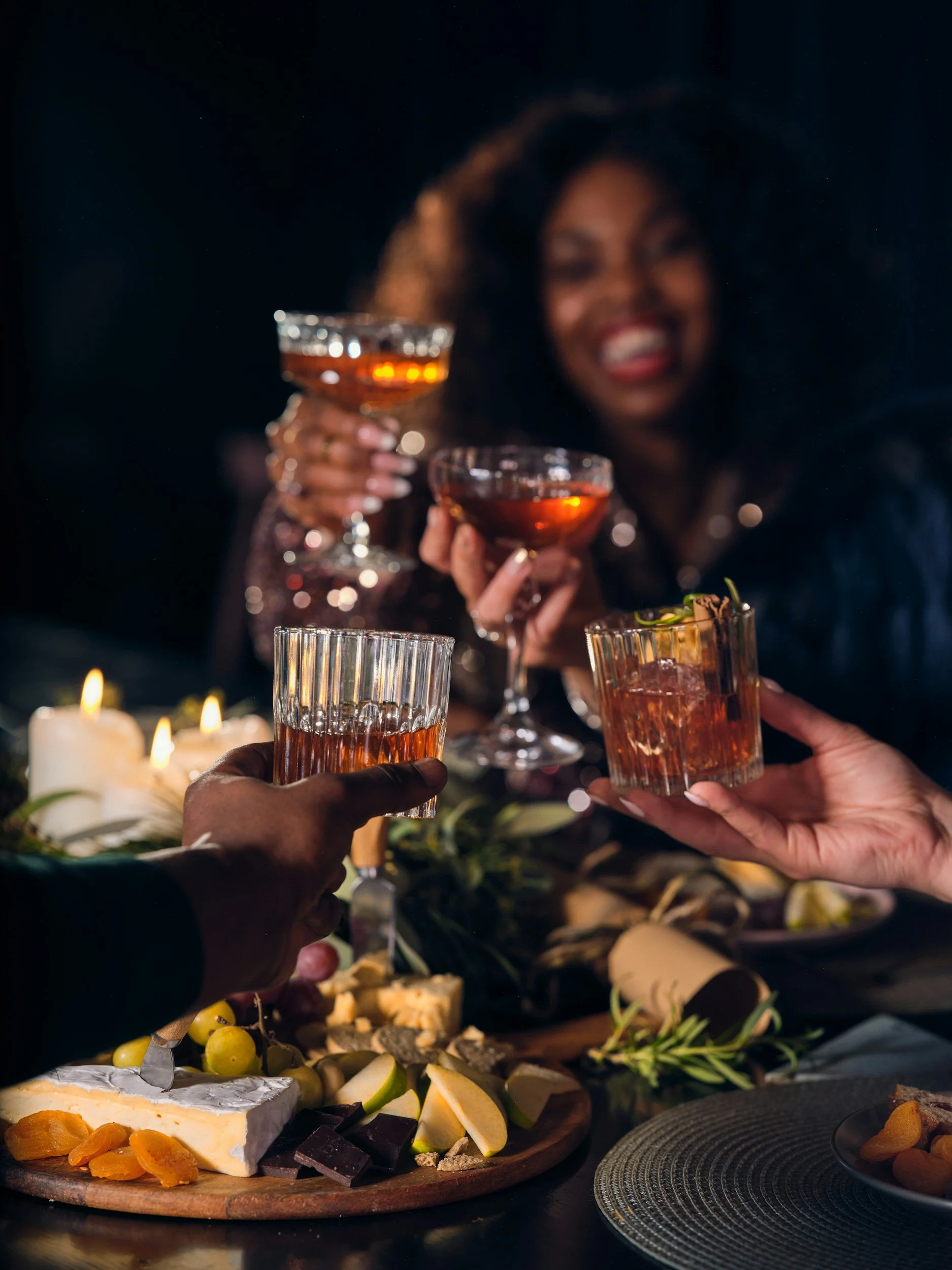 People clinking glasses of cocktails during a celebration with a cheese platter and candles on the table.