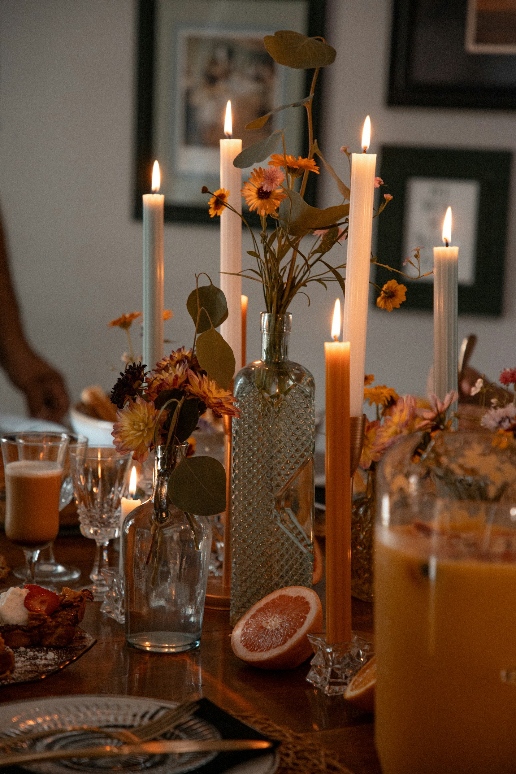 A dining table decorated with lit candles, floral arrangements in glass vases, and a halved grapefruit, creating a warm, cozy atmosphere.