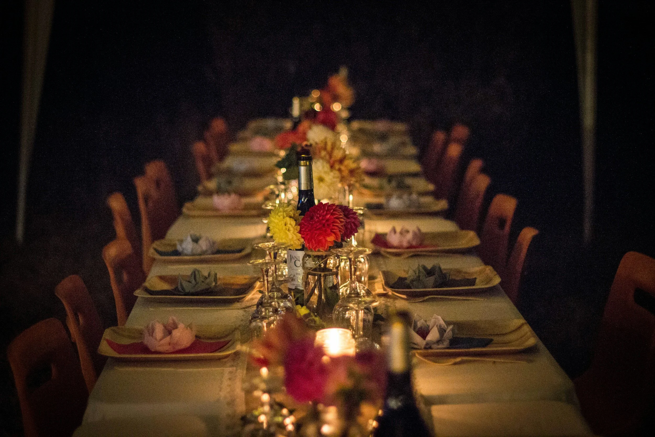 A long banquet table set for a dinner with floral centerpieces, wine bottles, and place settings, illuminated by candlelight in a dark room.