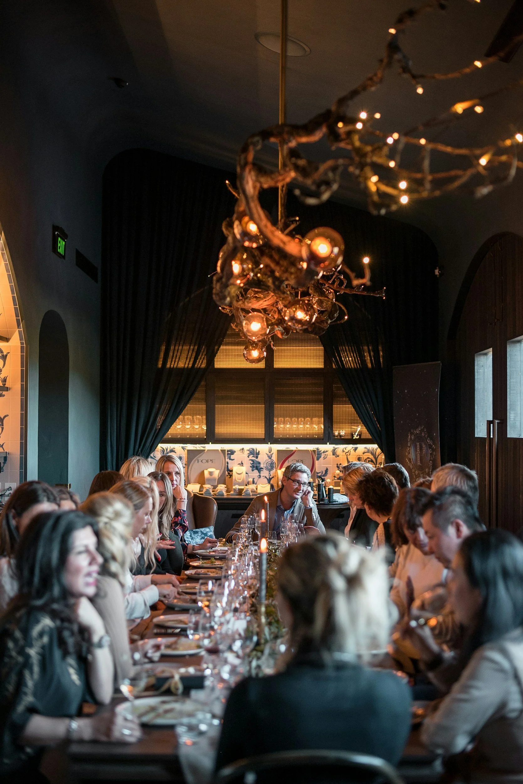 A large group of people dining at a long table in a dimly lit restaurant with dark curtains and a decorative chandelier hanging from the ceiling.