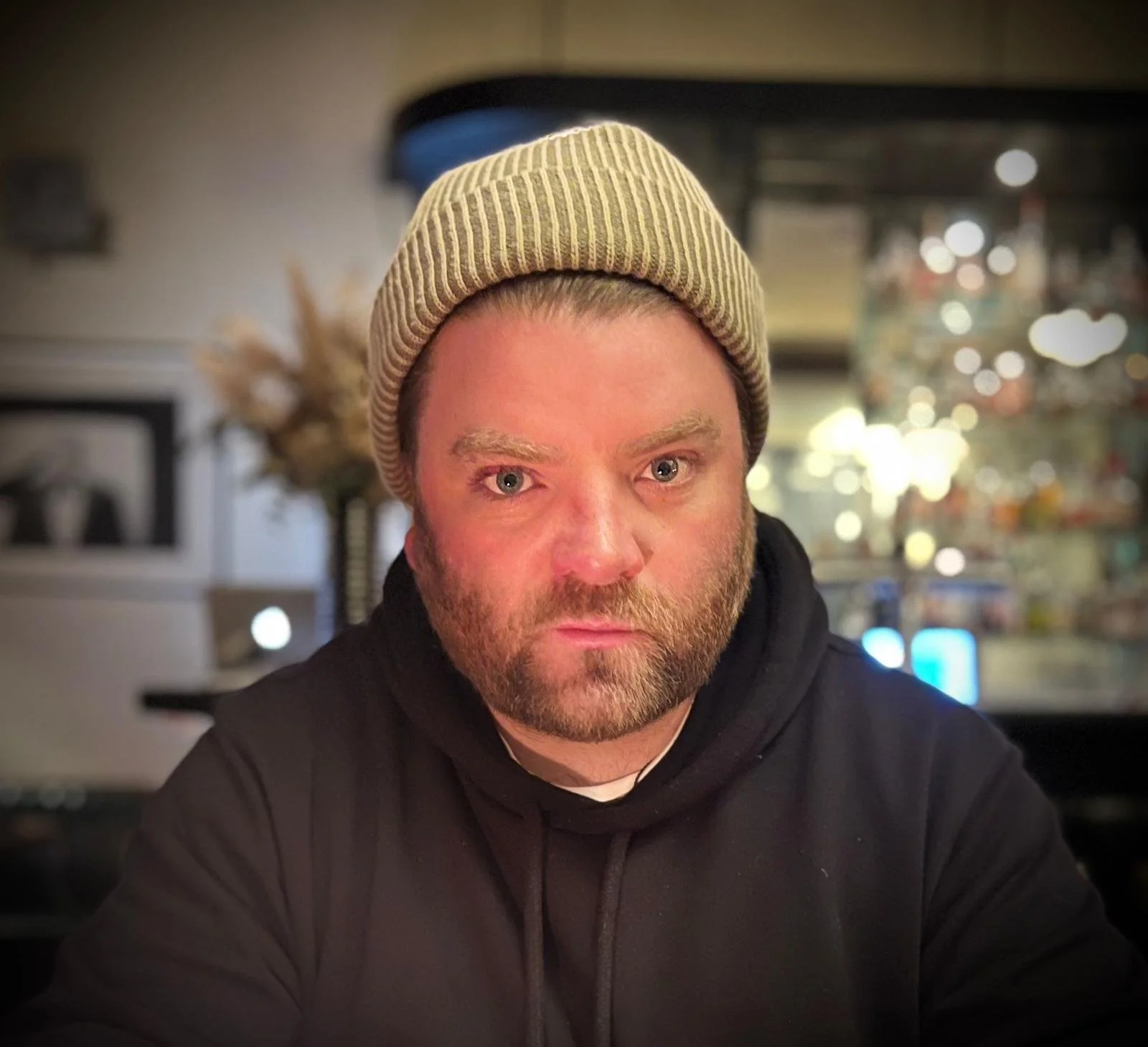 A man with a beard and light-colored eyebrows wearing a striped beige knit beanie and a black hoodie, sitting indoors with a blurred background of home decor and bokeh lights.
