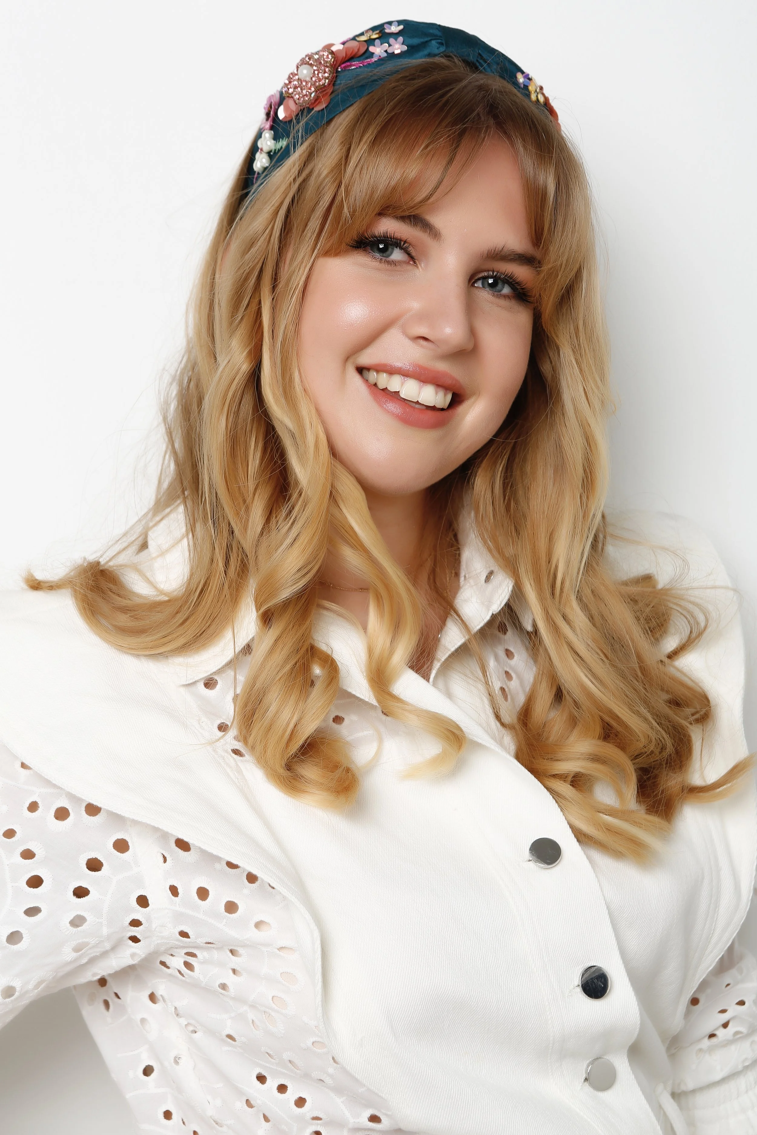 A young woman with long, wavy blonde hair, blue eyes, and light skin, smiling and looking to the side. She is wearing a navy blue headscarf decorated with colorful floral and pearl embellishments, and a white outfit with eyelet embroidery and silver buttons.