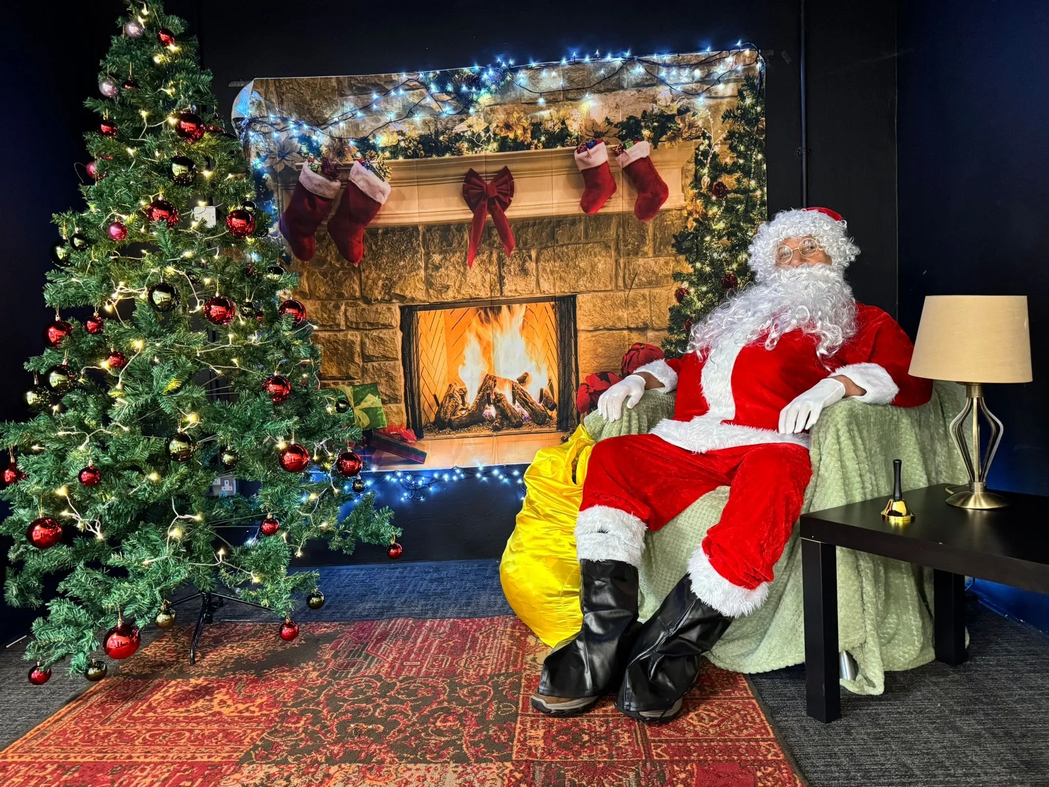 Santa Claus sitting in a green armchair in front of a fireplace decorated with Christmas stockings and lights, next to a decorated Christmas tree with red and gold ornaments.