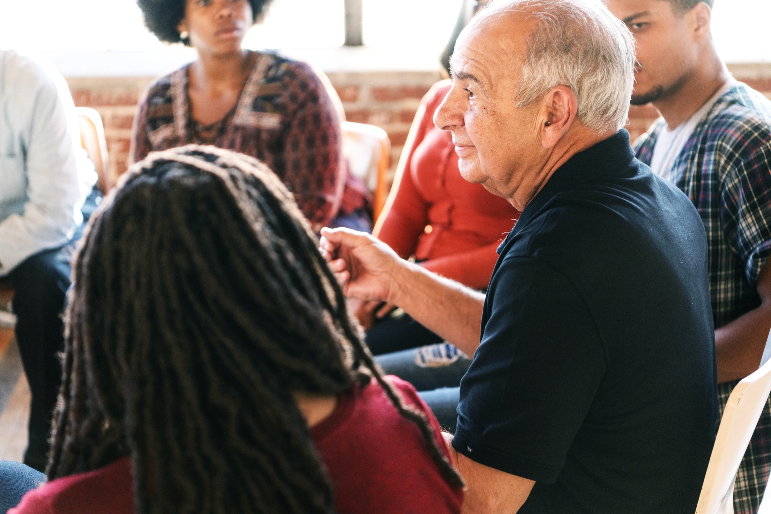An elderly man with gray hair talking during a discussion in a group setting, with diverse young adults seated around him.