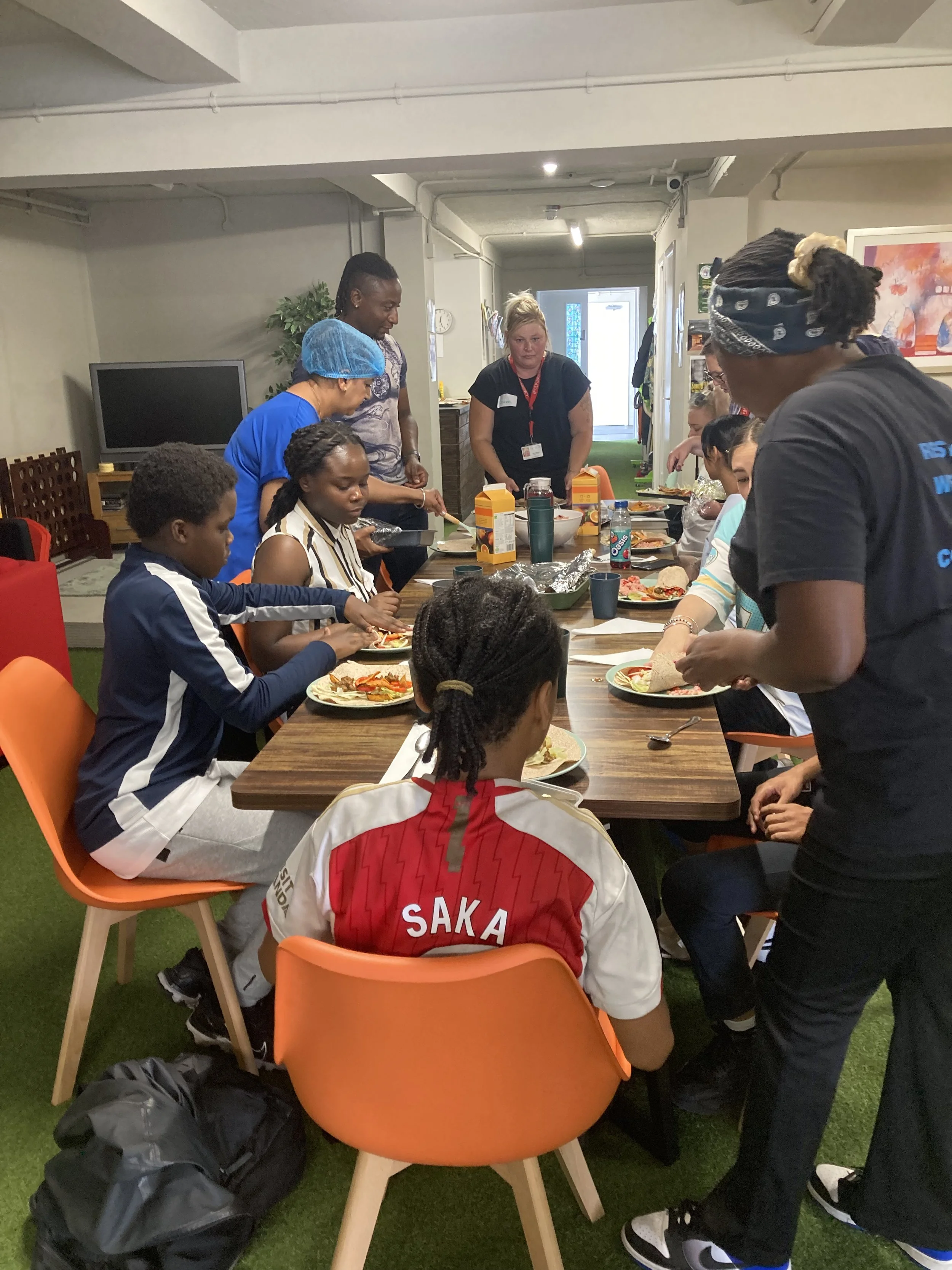 A group of adults and children gathered around a table in a room, ready to eat a meal, with various food items, drinks, and utensils on the table.