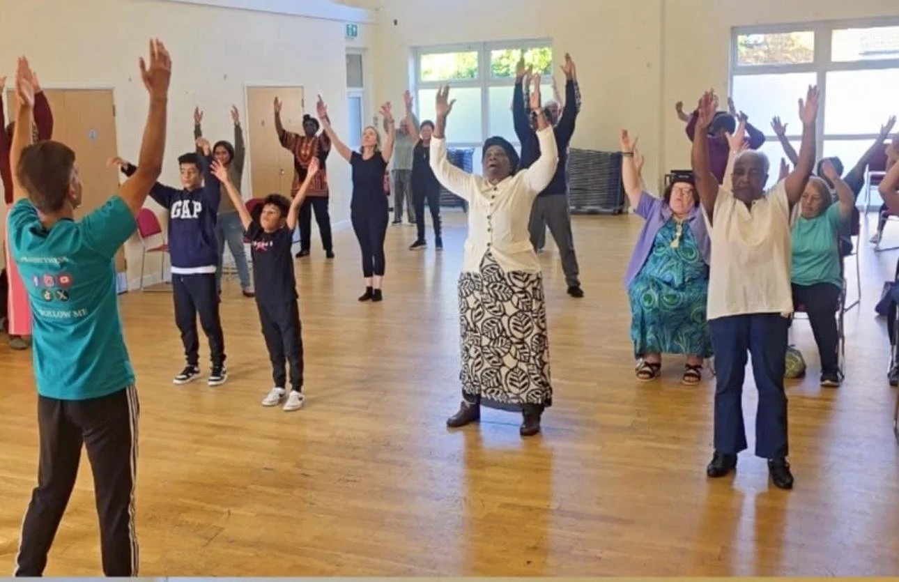 A diverse group of people, including children and elderly, participating in a dance or exercise class in a community hall, following instructor's movements with arms raised.
