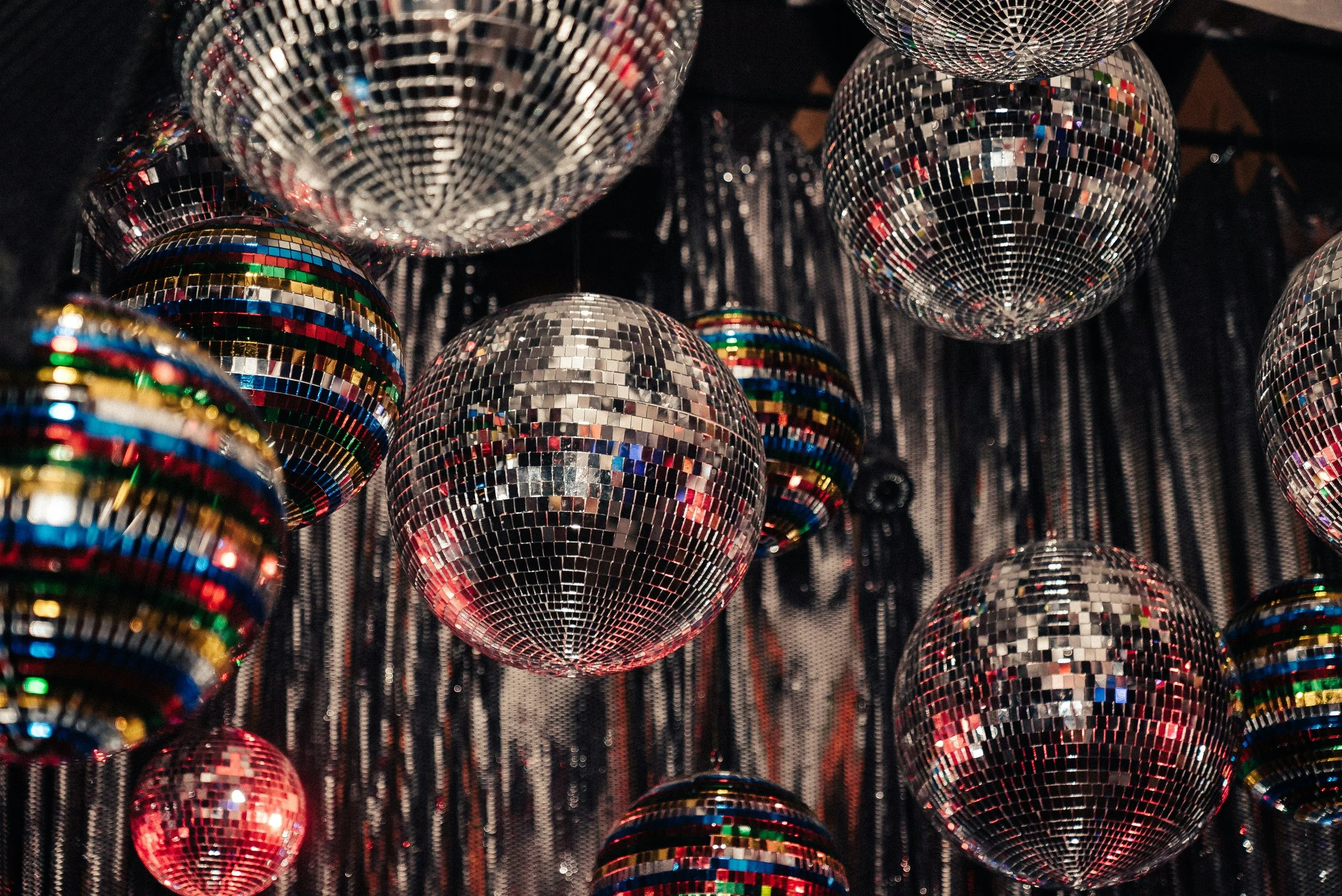 Multiple disco balls hanging against a black background with shimmering silver fringe. The disco balls are reflecting colorful lights.