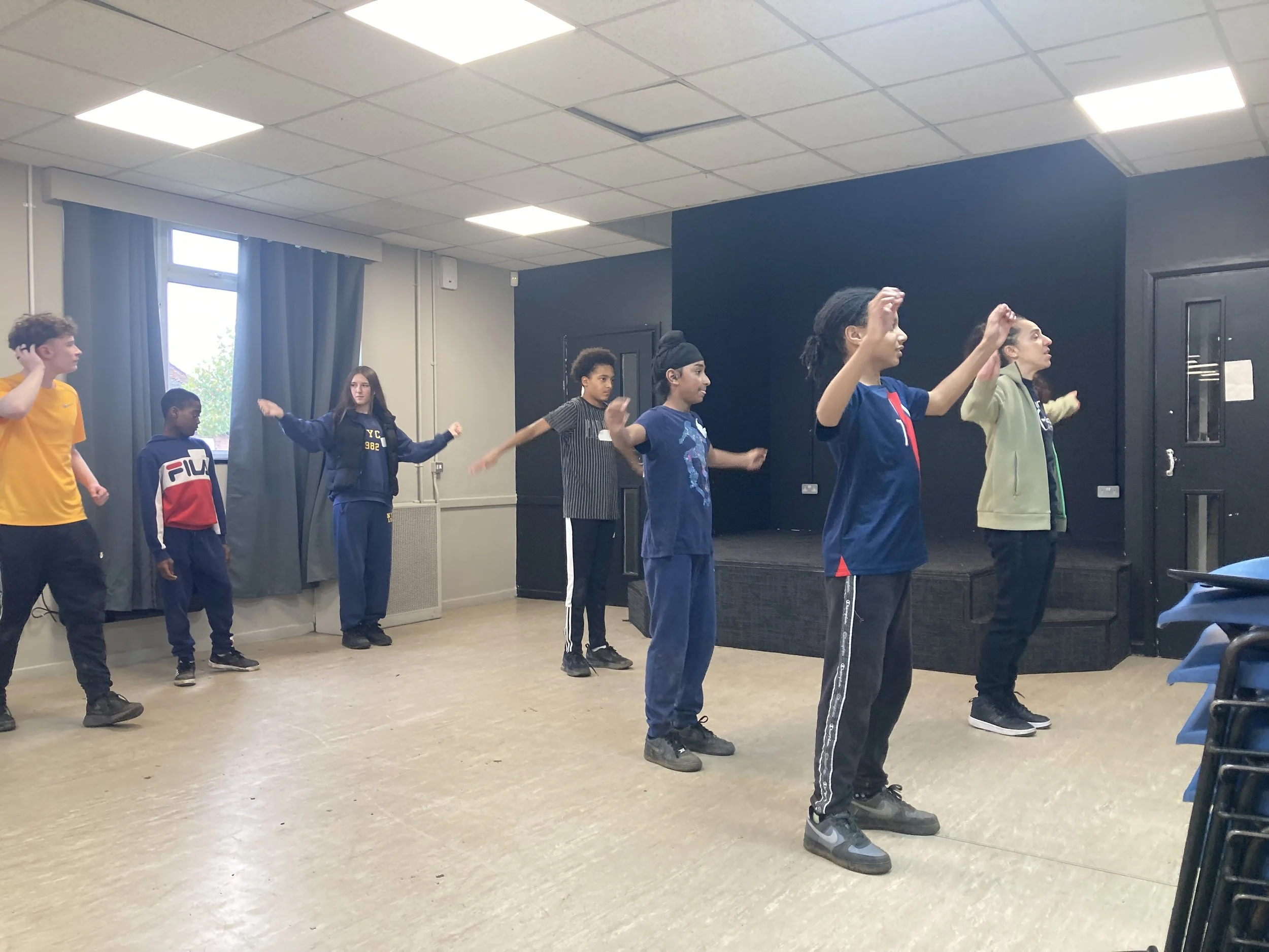A group of children practicing dance or theater moves on a stage in a classroom or studio setting.