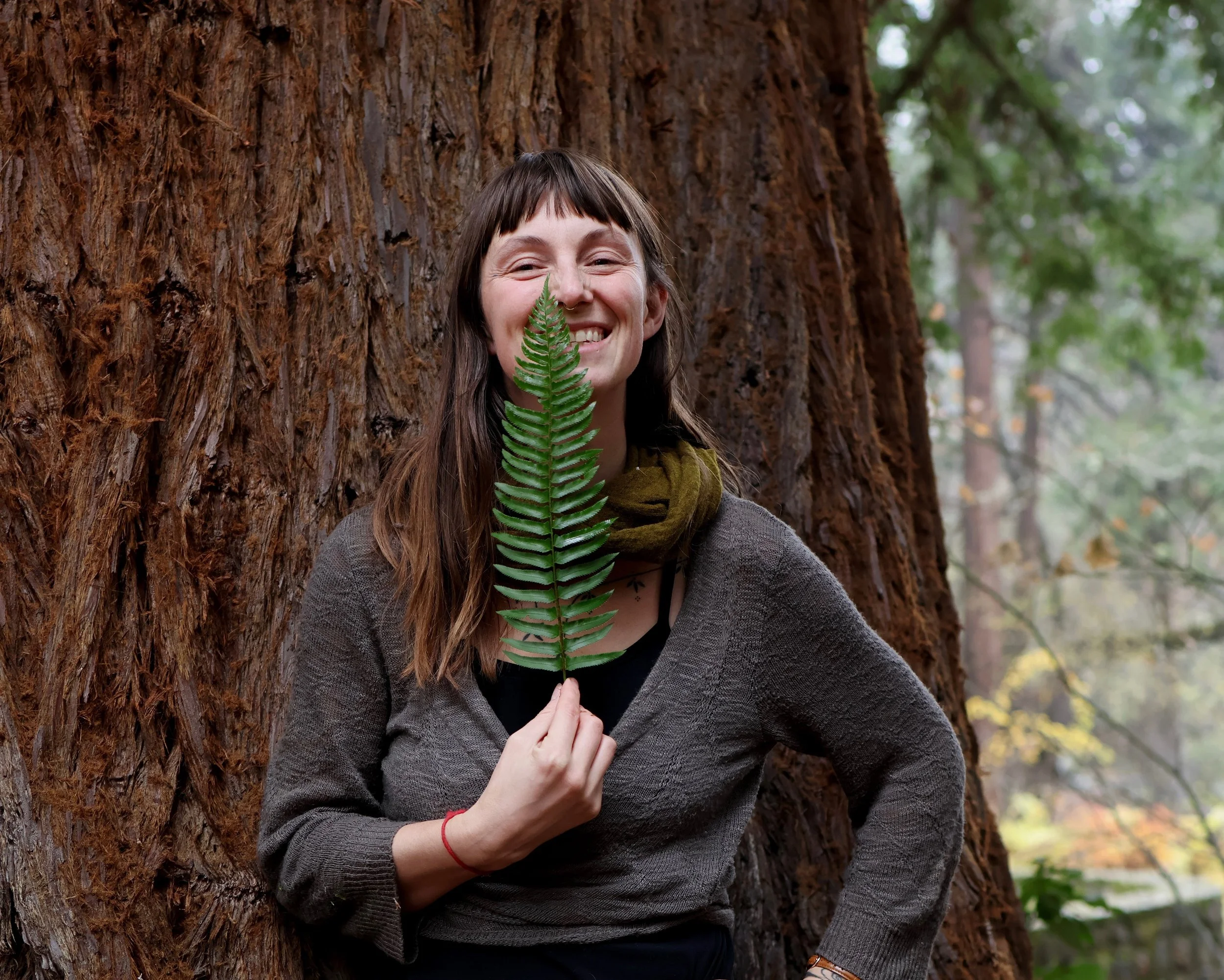 Fallyn McLeod stands in front of a redwood tree, holding a fern leaf to her face and smiling