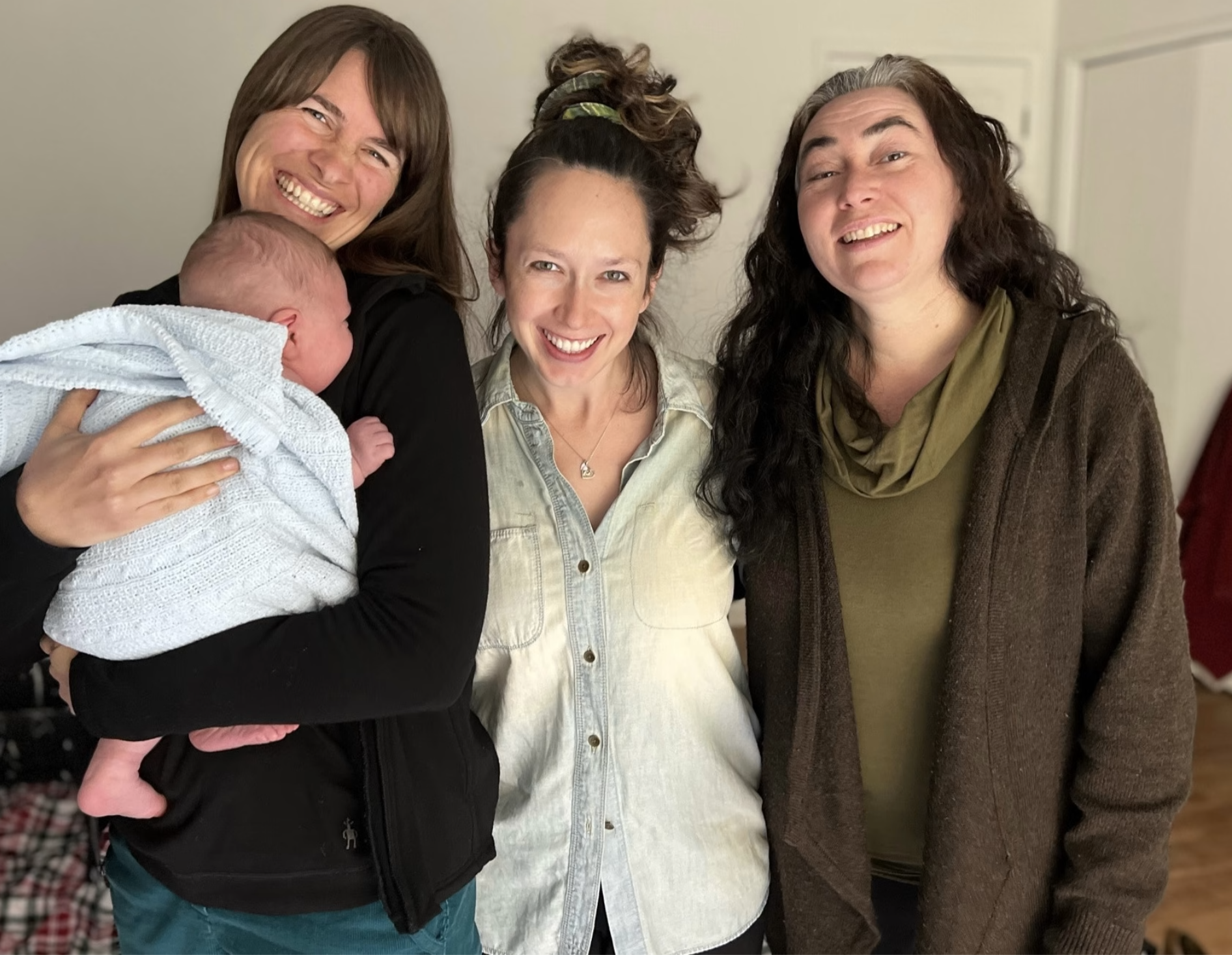 Four women and one baby smiling for a photo indoors. One woman is holding the baby wrapped in a white blanket, all are smiling and happy.