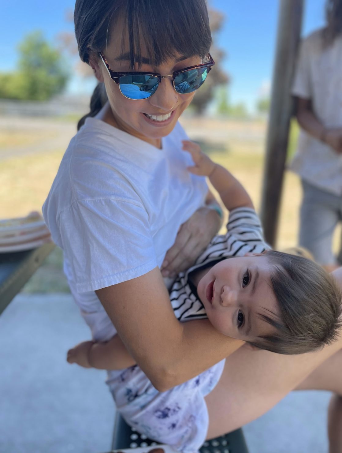 A woman wearing sunglasses and a white shirt is holding a young boy with brown hair and a striped shirt. They are outdoors on a sunny day, smiling and enjoying each other's company.
