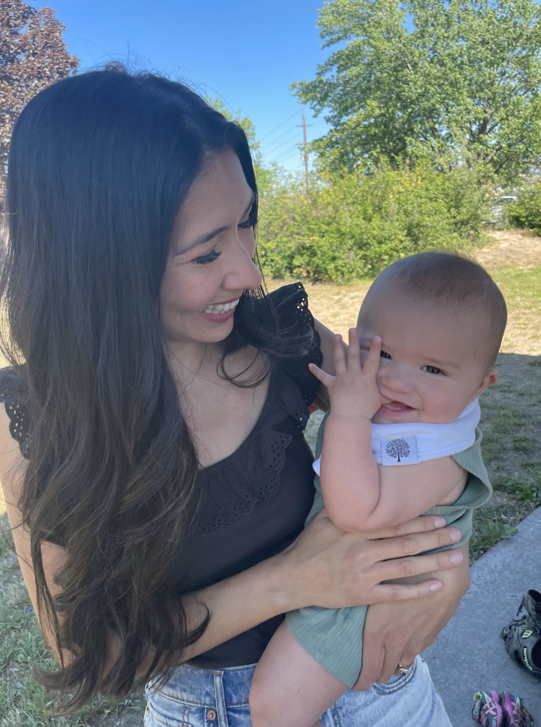 A woman holding a baby outdoors on a sunny day, both smiling and looking at each other.