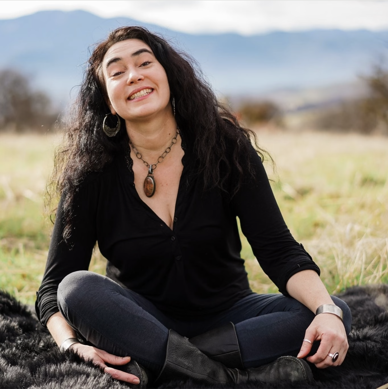 Veege Ruediger sits cross-legged in a field with mountains in the backdrop