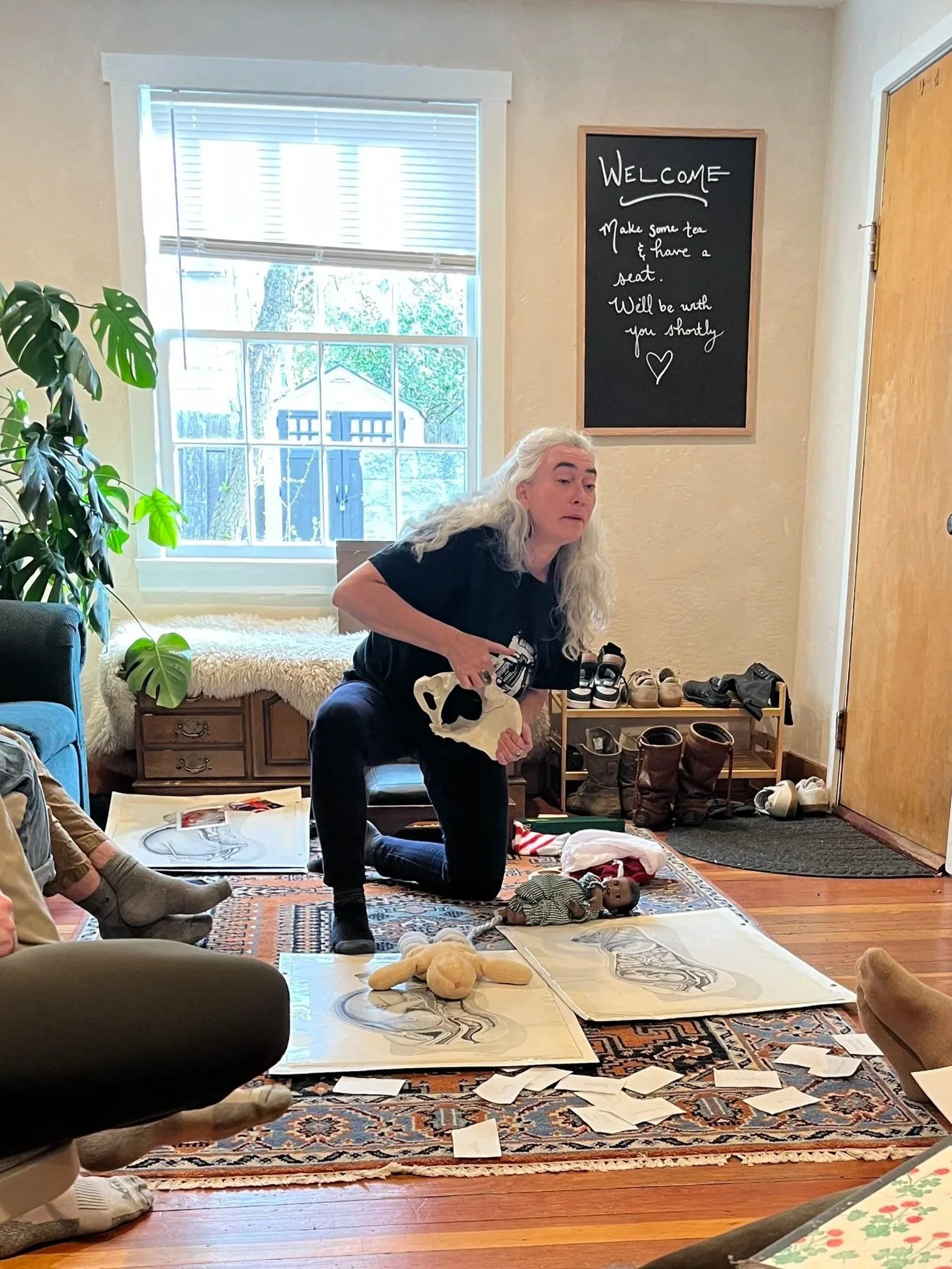 A woman with long white hair kneeling on a patterned rug in a room with a large window, surrounded by toys, shoes, and papers, holding a pair of black shoes.