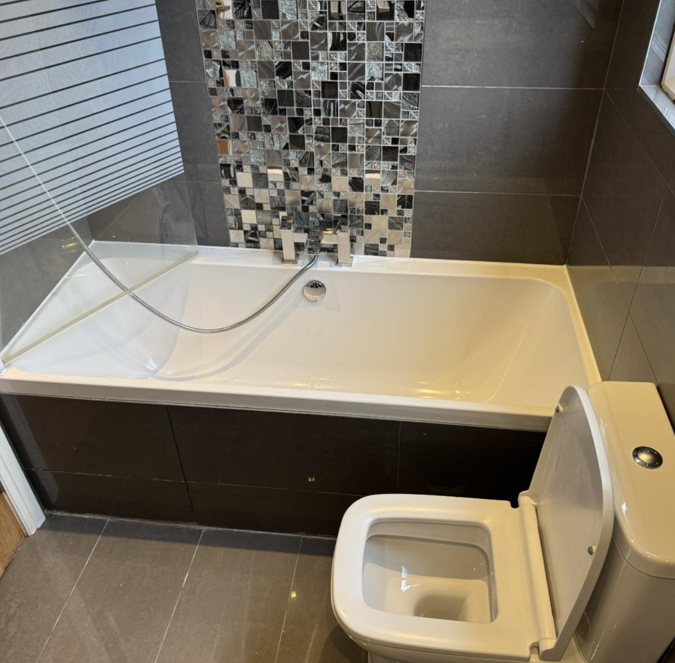 Bathroom with a white bathtub, black tile panels, mosaic tile accent wall, and a section of frosted glass.