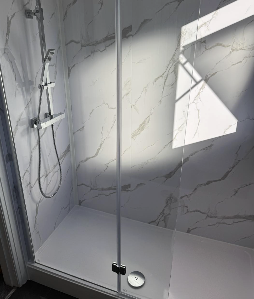 Empty walk-in shower with white marble walls and a glass door, sunlight casting shadow on the wall.