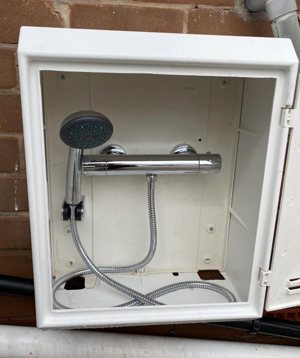 Uninstalled outdoor shower with a handheld showerhead and flexible hose mounted inside a white enclosure on an exterior brick wall.