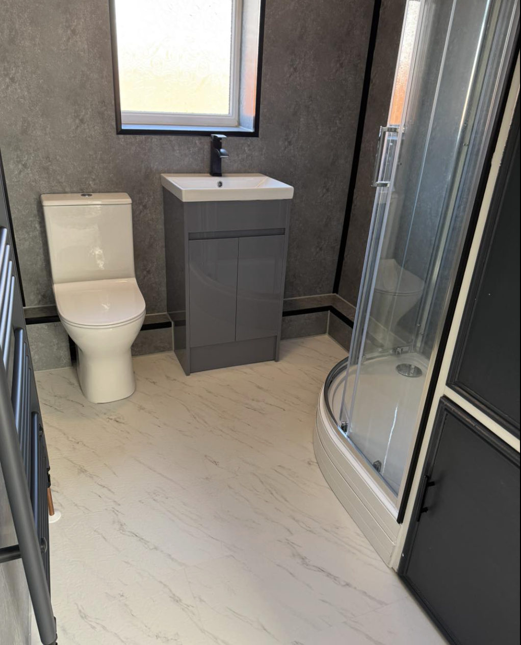 A modern bathroom with a white toilet, a small gray vanity with a sink and black faucet, a frosted window, and a glass corner shower with sliding doors, set against textured gray walls and a light-colored marble-patterned floor.
