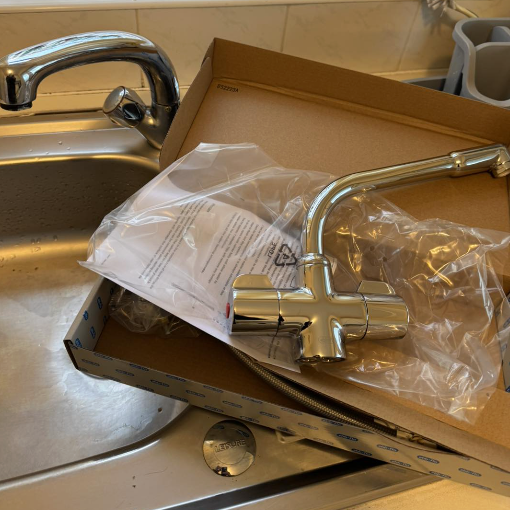 New chrome kitchen faucet placed on a cardboard box next to a kitchen sink, with a plastic bag and instruction sheet, in a kitchen setting.