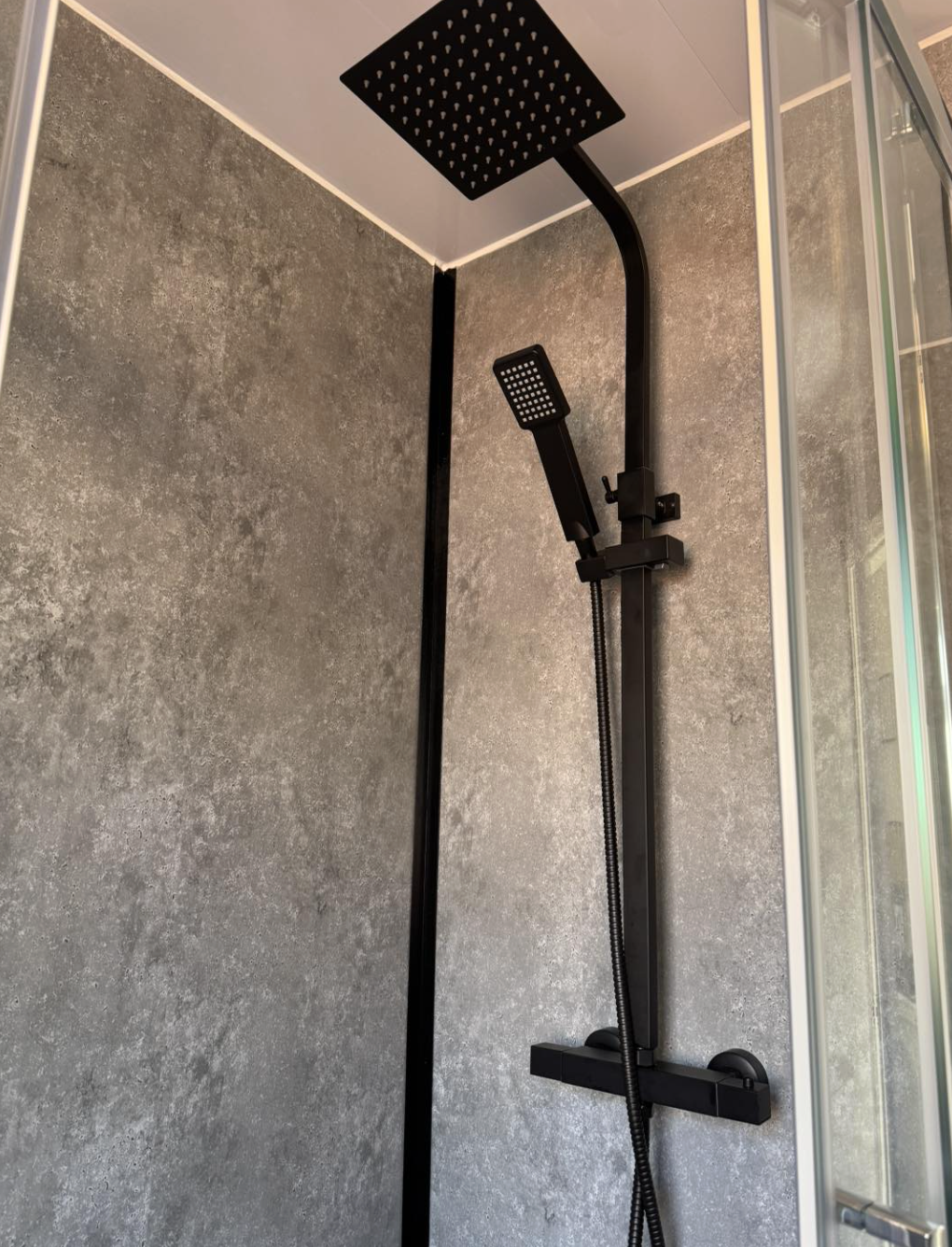 Black shower set with a rain showerhead and a handheld showerhead in a modern shower enclosure with gray textured wall and glass door.