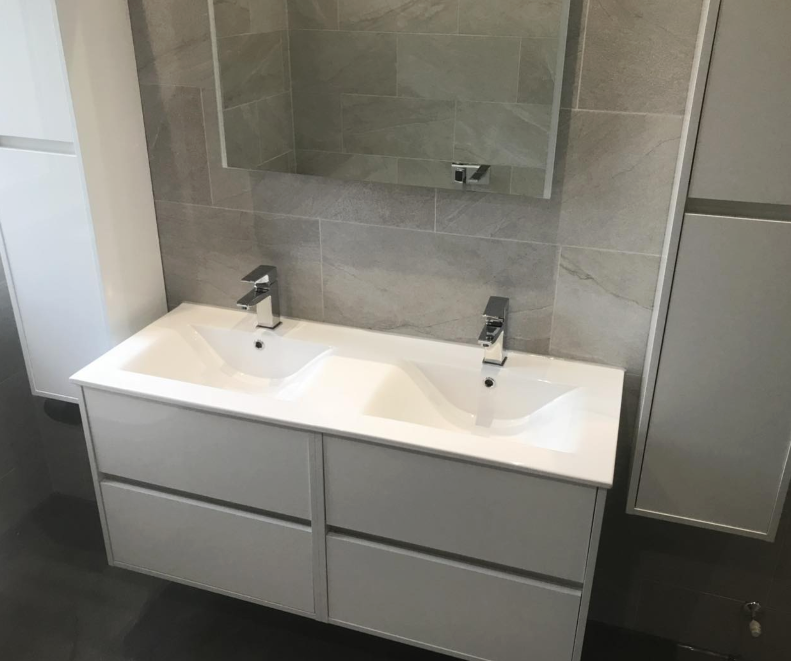 Modern bathroom sink vanity with dual sinks, chrome faucets, a large mirror above, and gray tiled wall behind.