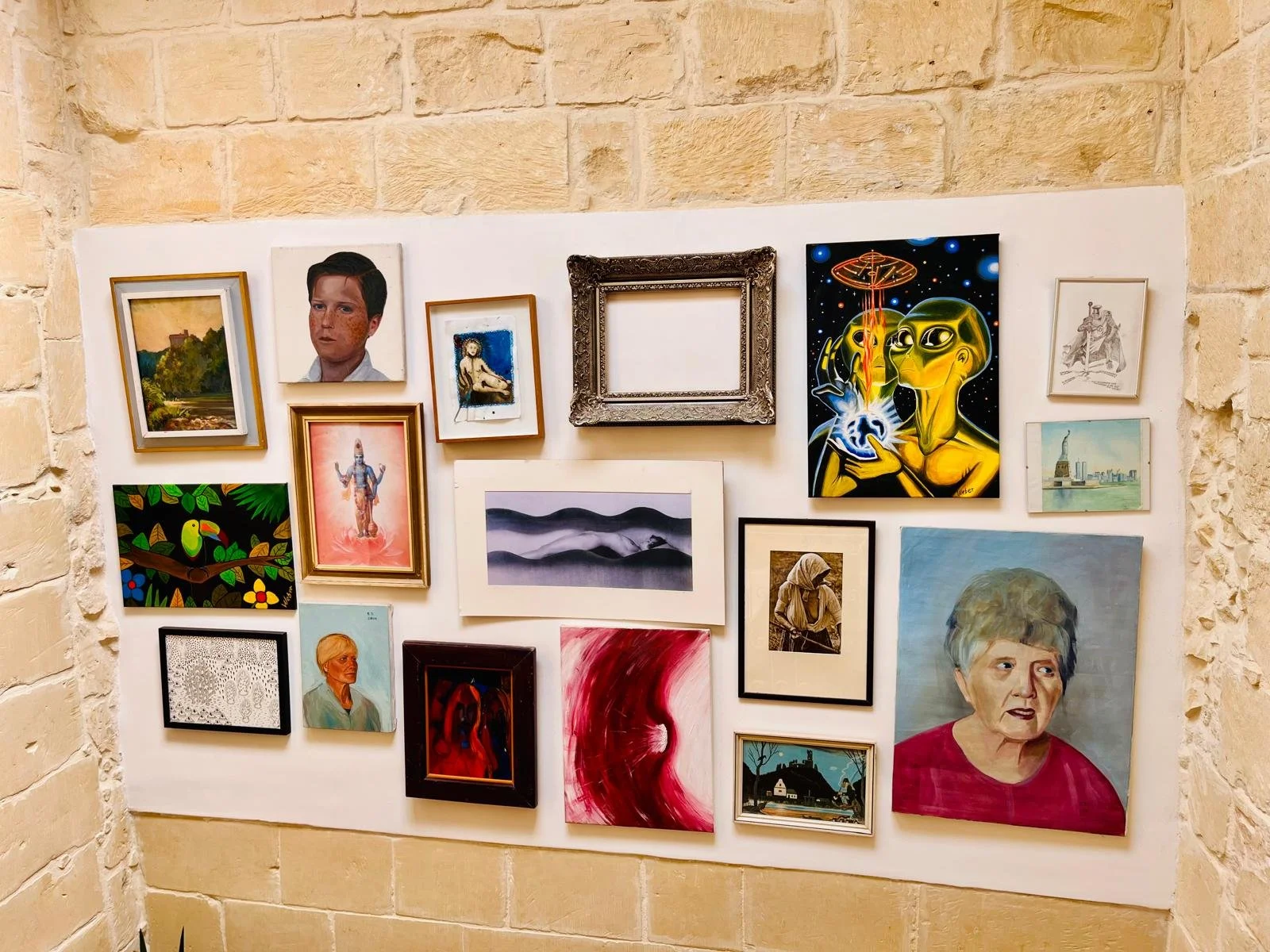 A wall display of various framed artworks, including portraits, landscapes, abstract pieces, and imaginative illustrations, arranged on a white wall within a room with beige brick walls.