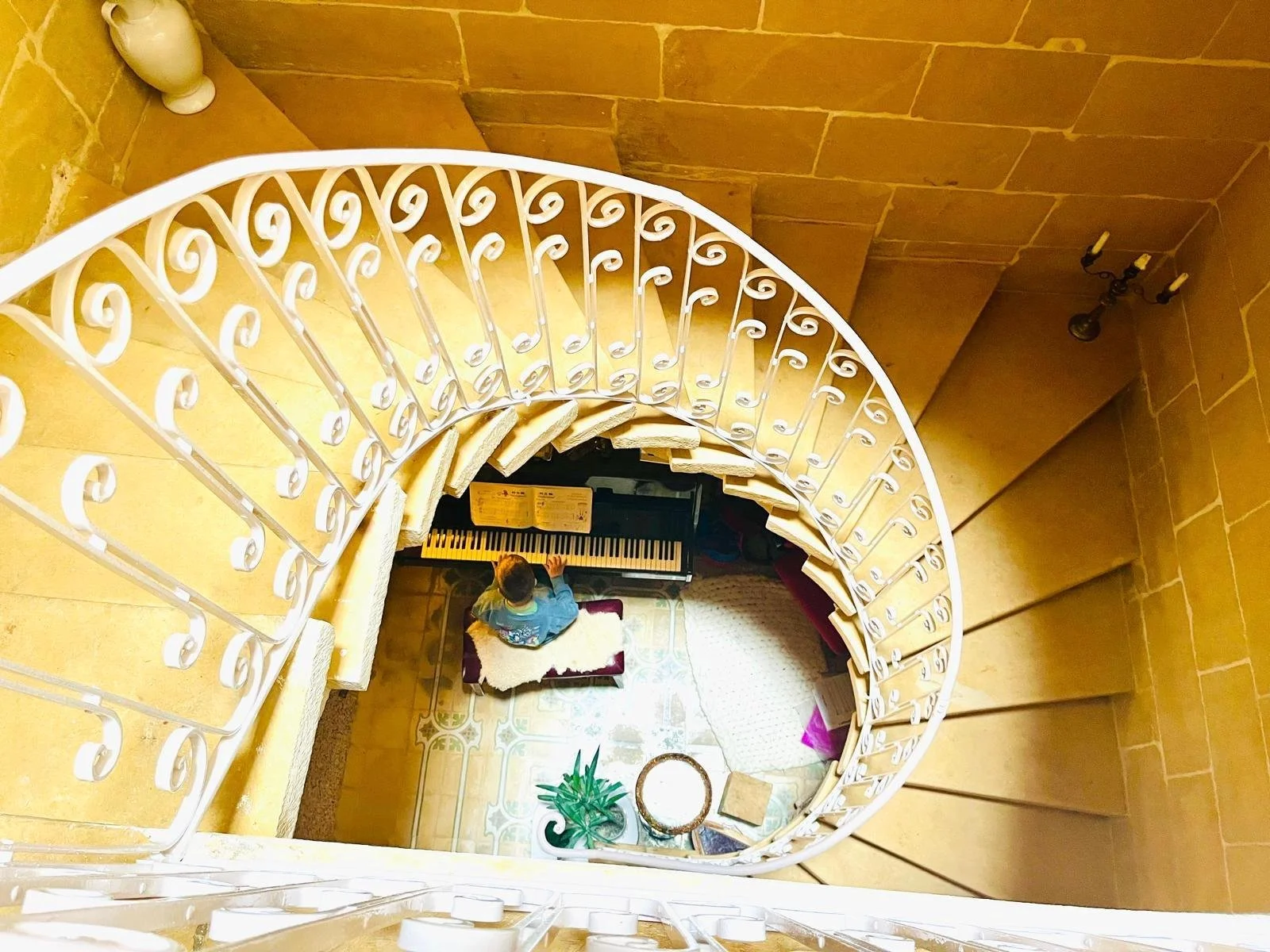 Looking down a spiral staircase with a black and white piano at the bottom, a person playing it, decorative plants, and vintage wall lamps.