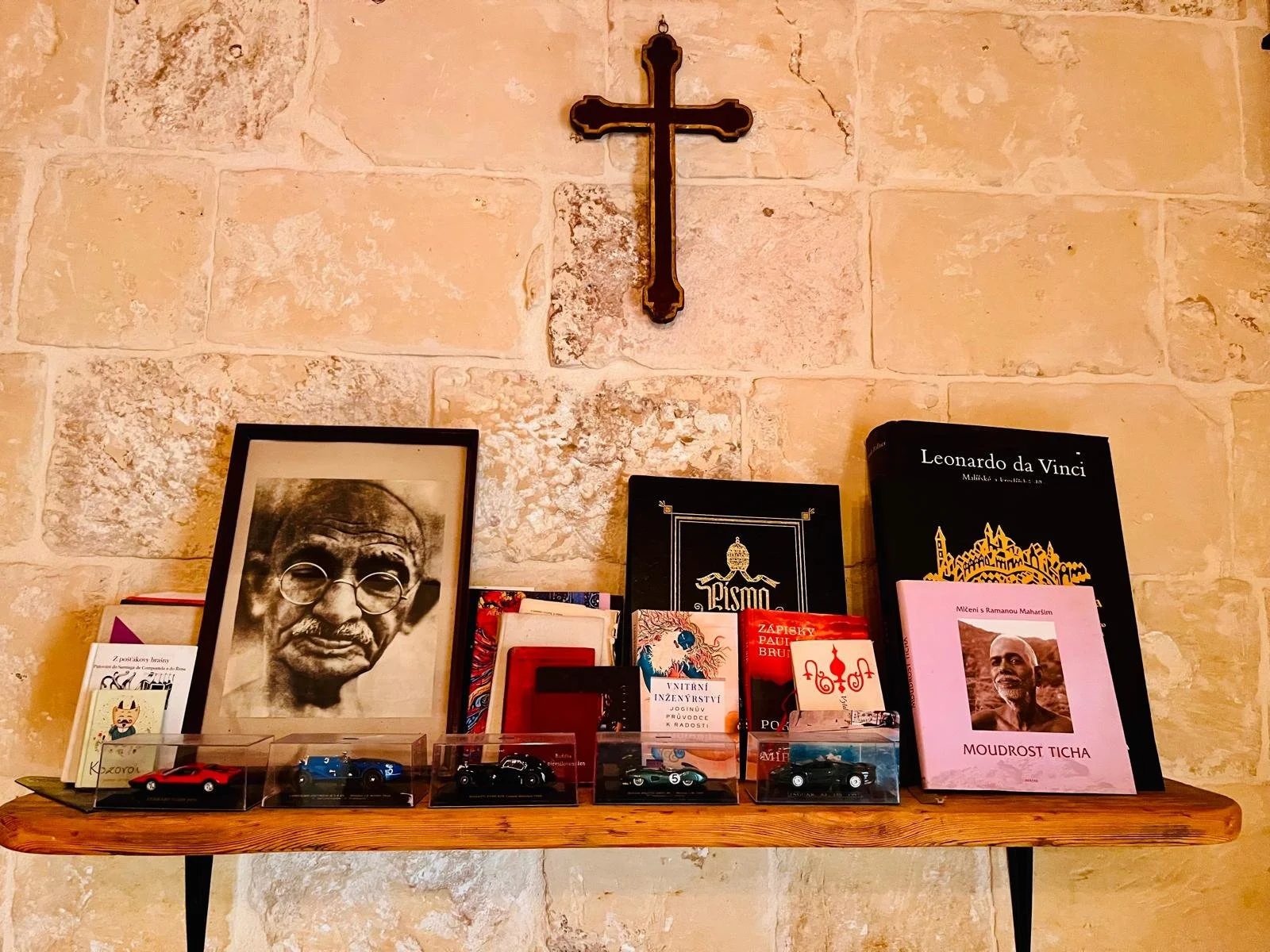 A wooden shelf against a stone wall with a black cross mounted above it. The shelf holds a framed black and white picture of Mahatma Gandhi, several books including titles on Leonardo da Vinci and other topics, a pink book titled "Moudrost Ticha" fea