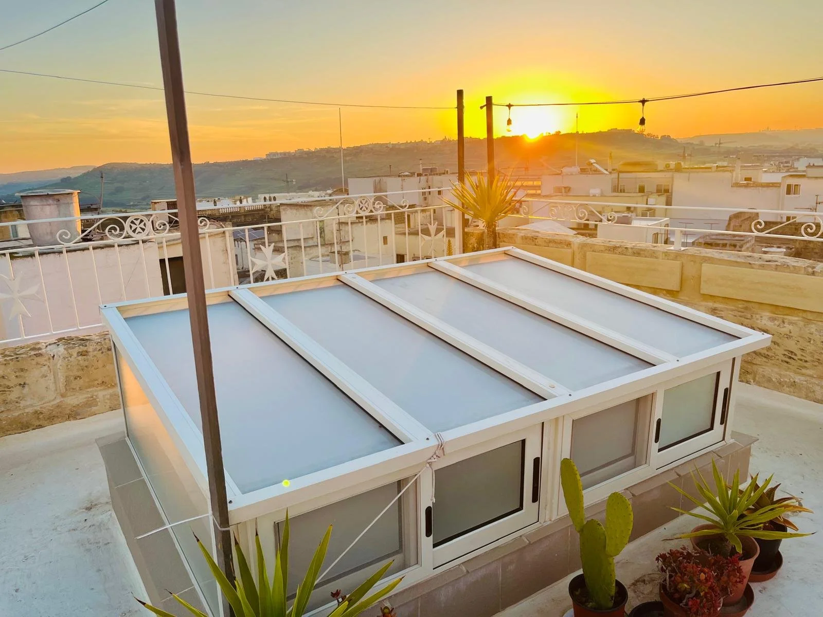 Sunset view from a rooftop terrace with potted plants and a glass-covered structure, overlooking buildings and hills in the distance.