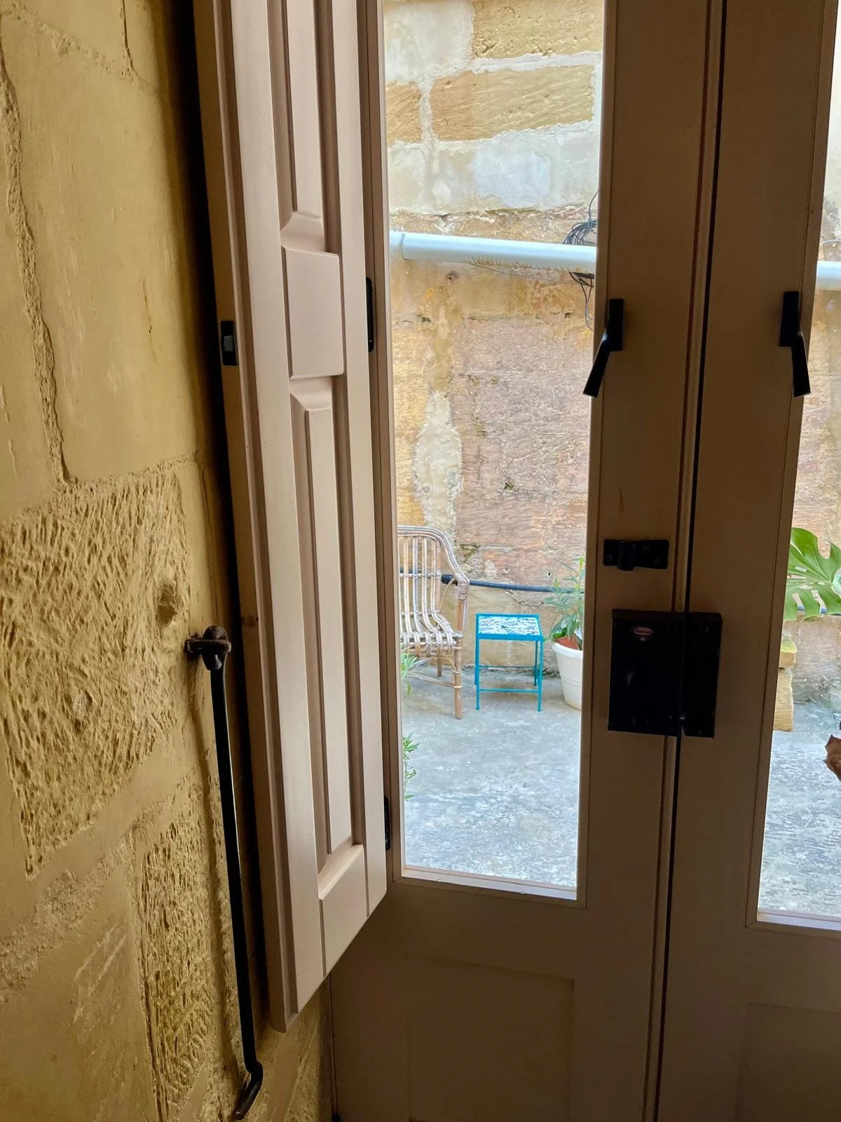 View of a small outdoor patio area through a glass door with a brass handle. The patio has a wooden chair, a small blue table, a potted plant, and a stone wall.